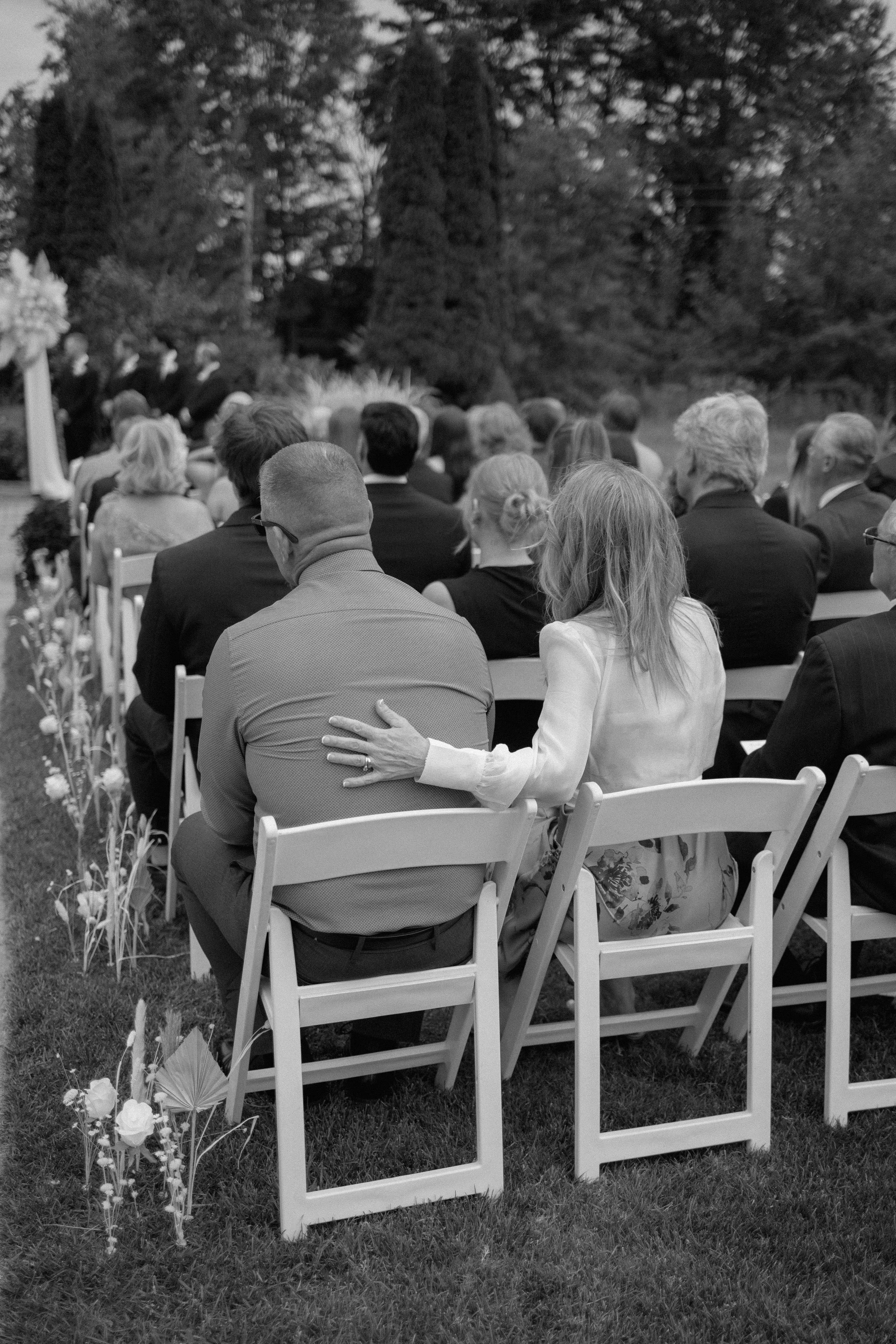 A black and white photo of a group of people seated outdoors on white chairs, attending a formal event such as a wedding or ceremony. The setting appears to be in a garden or park, with trees and flowers visible in the background.