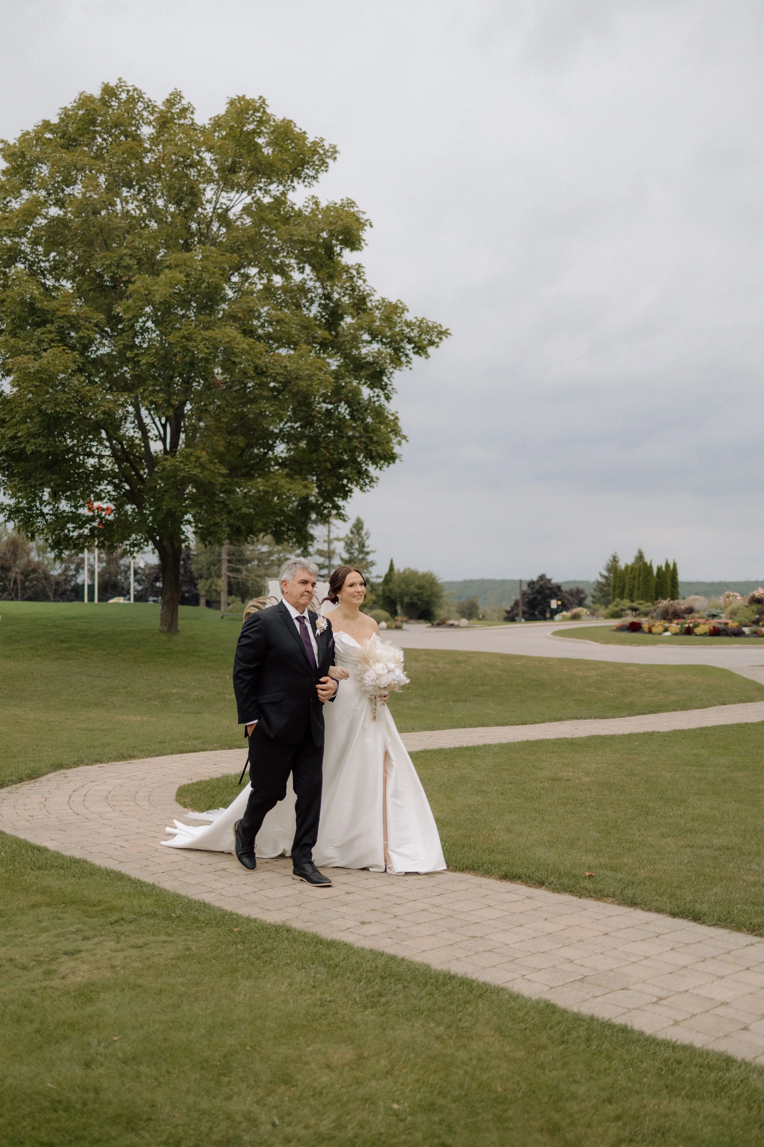 A bride and a man, possibly her father, walking together on a curved brick pathway in a park during daytime. The bride is wearing a white wedding gown and holding a bouquet, while the man is dressed in a dark suit with a boutonnière. The background f
