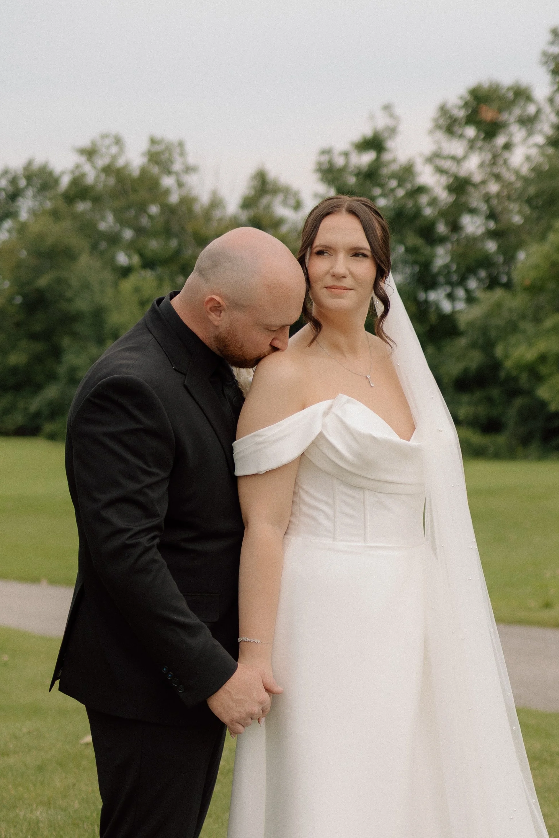A bride and groom holding hands outdoors with green trees in the background. The groom kissess the bride's shoulder, who looks to the side with a slight smile.