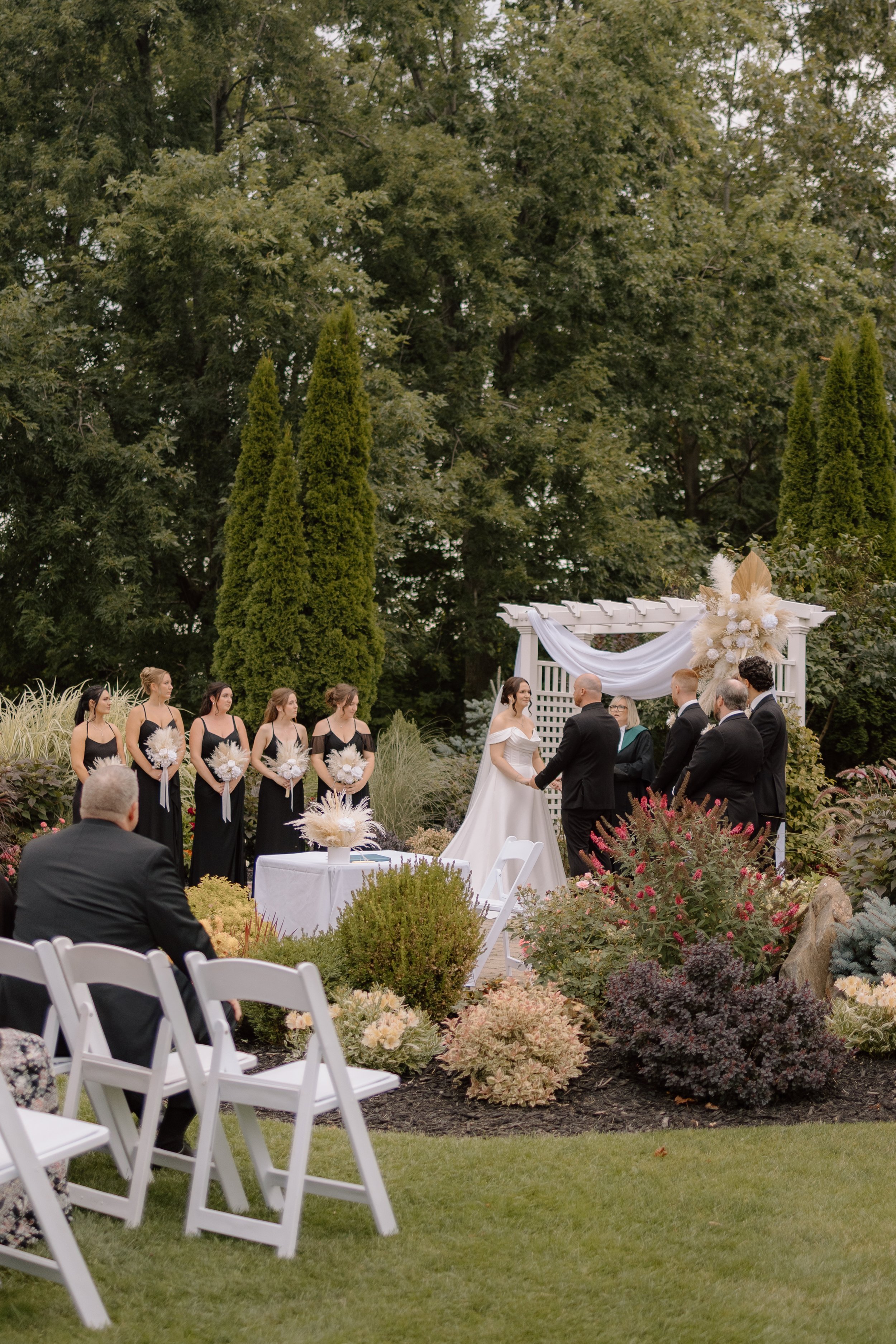 An outdoor wedding ceremony in a garden with married couple exchanging vows, surrounded by bridesmaids, groomsmen, guests, and floral arrangements, under a white pergola with draped fabric.