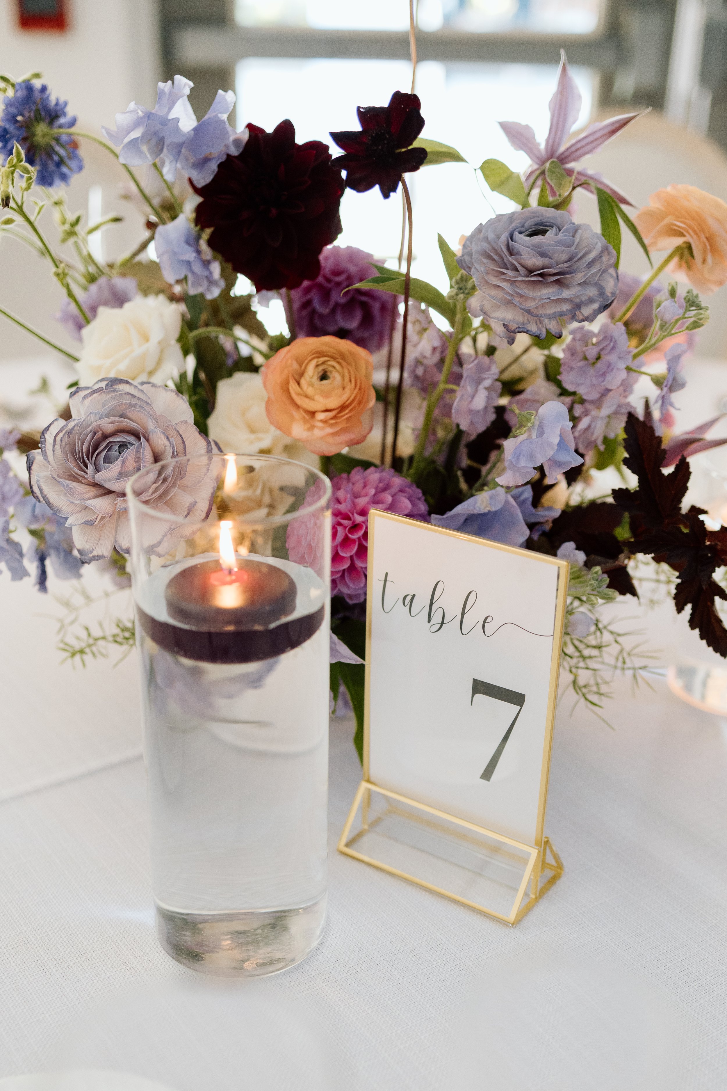 Vase with purple, pink, blue, white, and dark red flowers on a white table, candle in a glass holder, and a gold table number sign with the number 7.