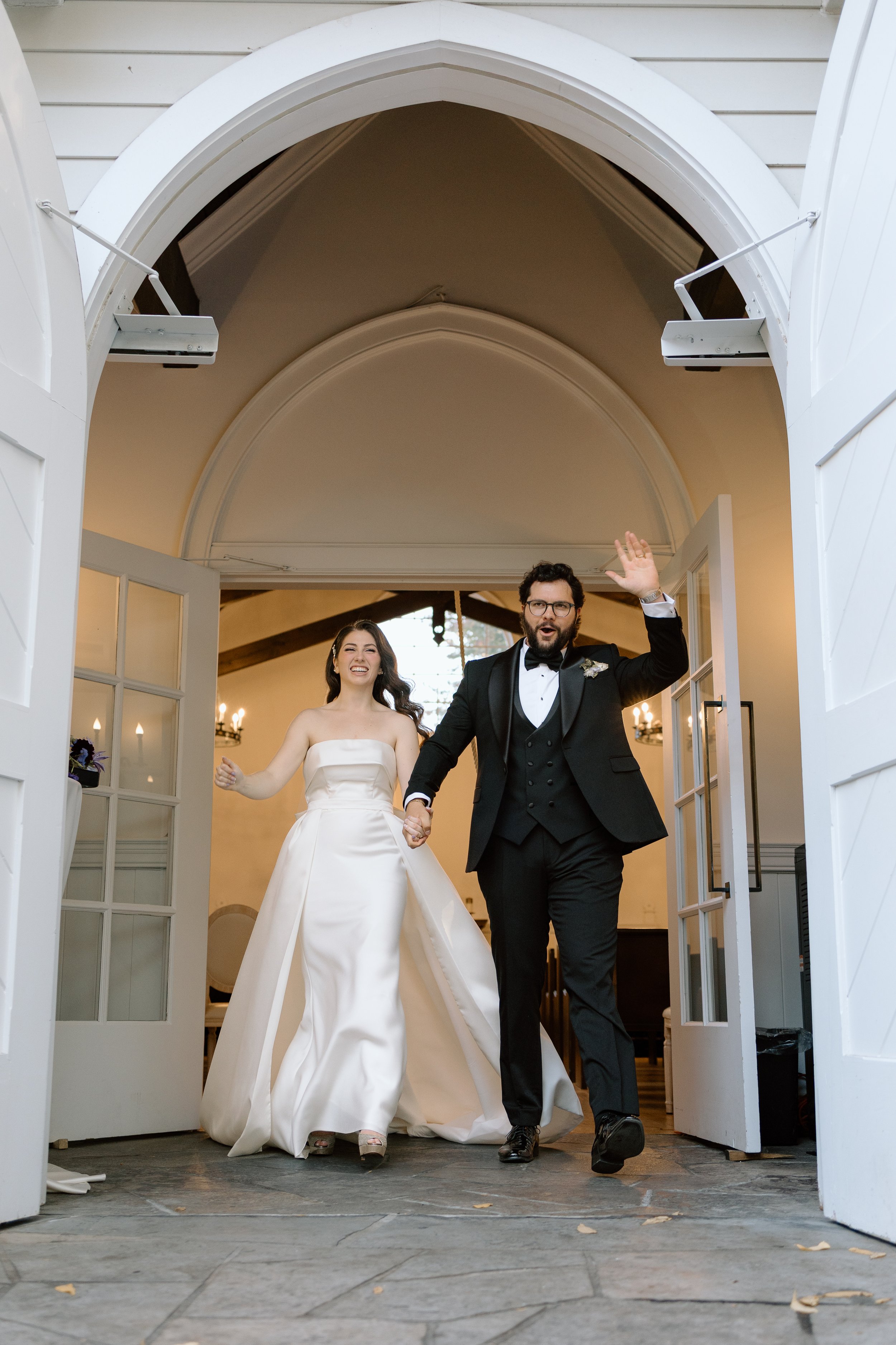 A bride and groom holding hands and walking through a set of open white doors at their wedding reception, smiling and celebrating.