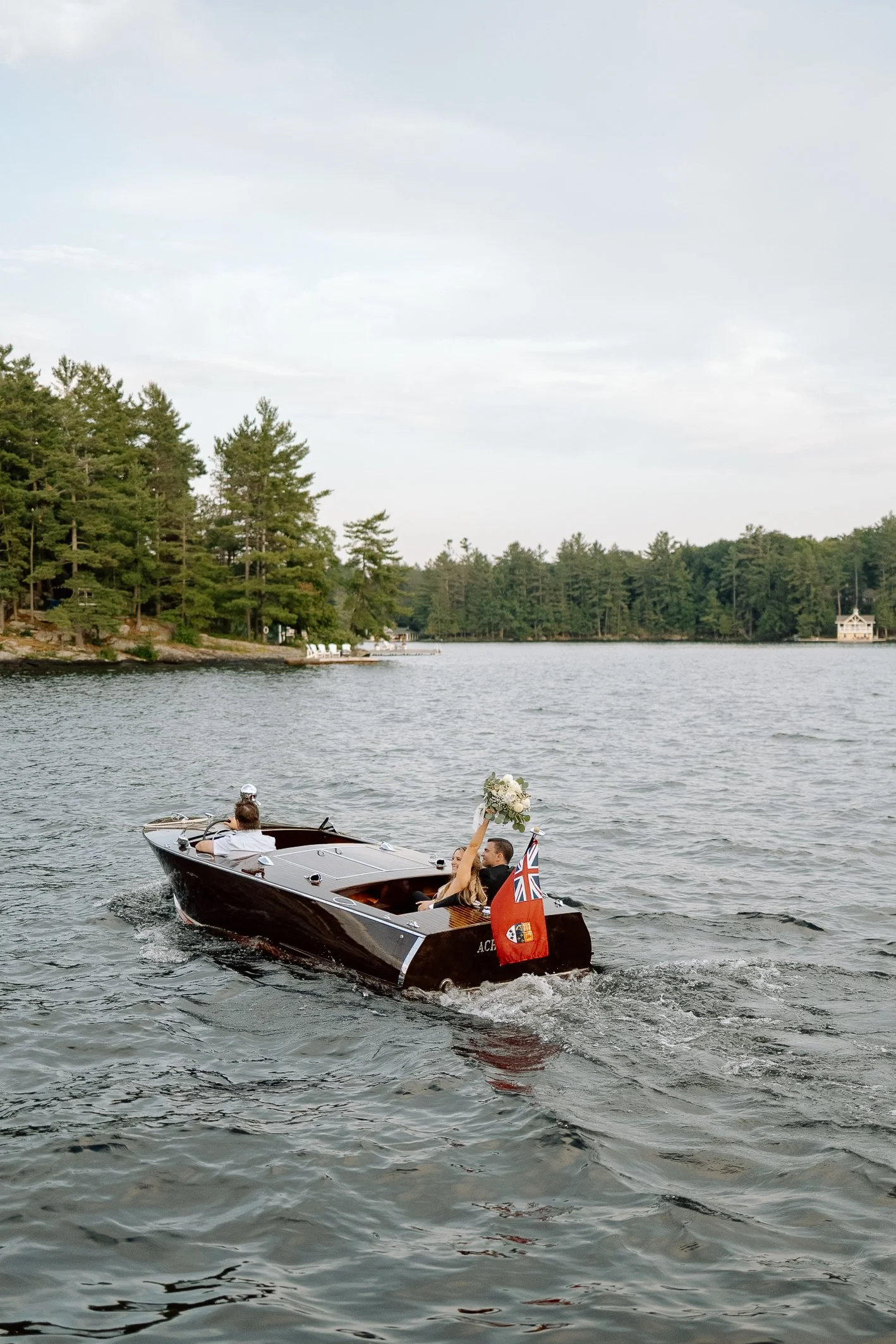 A couple is celebrating their wedding on a small motorboat on a lake. The bride is holding a bouquet of white flowers, and the groom is holding a Union Jack flag. The boat is black with white trim, and the water is calm with gentle ripples. In the ba