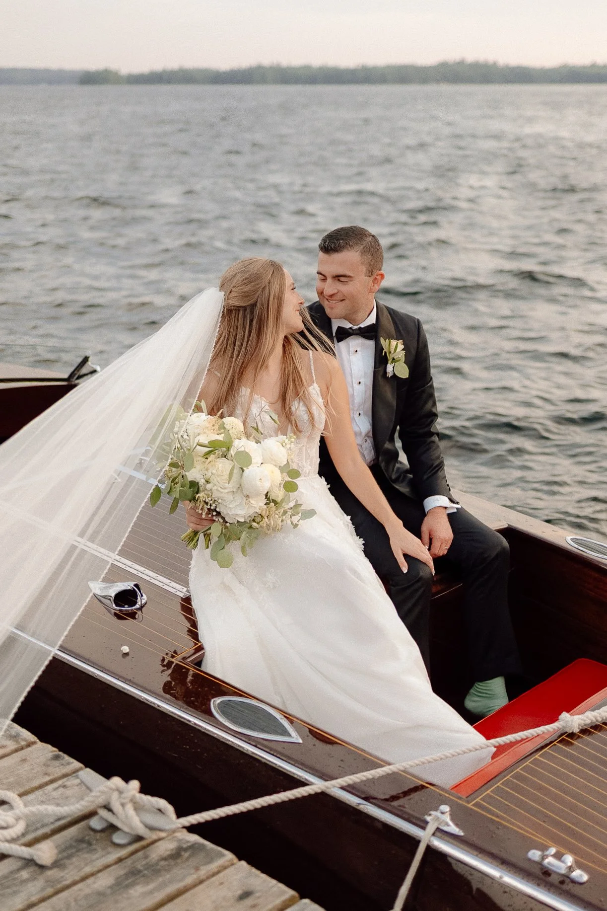 A bride and groom sitting together on a boat by the water, smiling and looking at each other, with the bride holding a bouquet of white flowers.