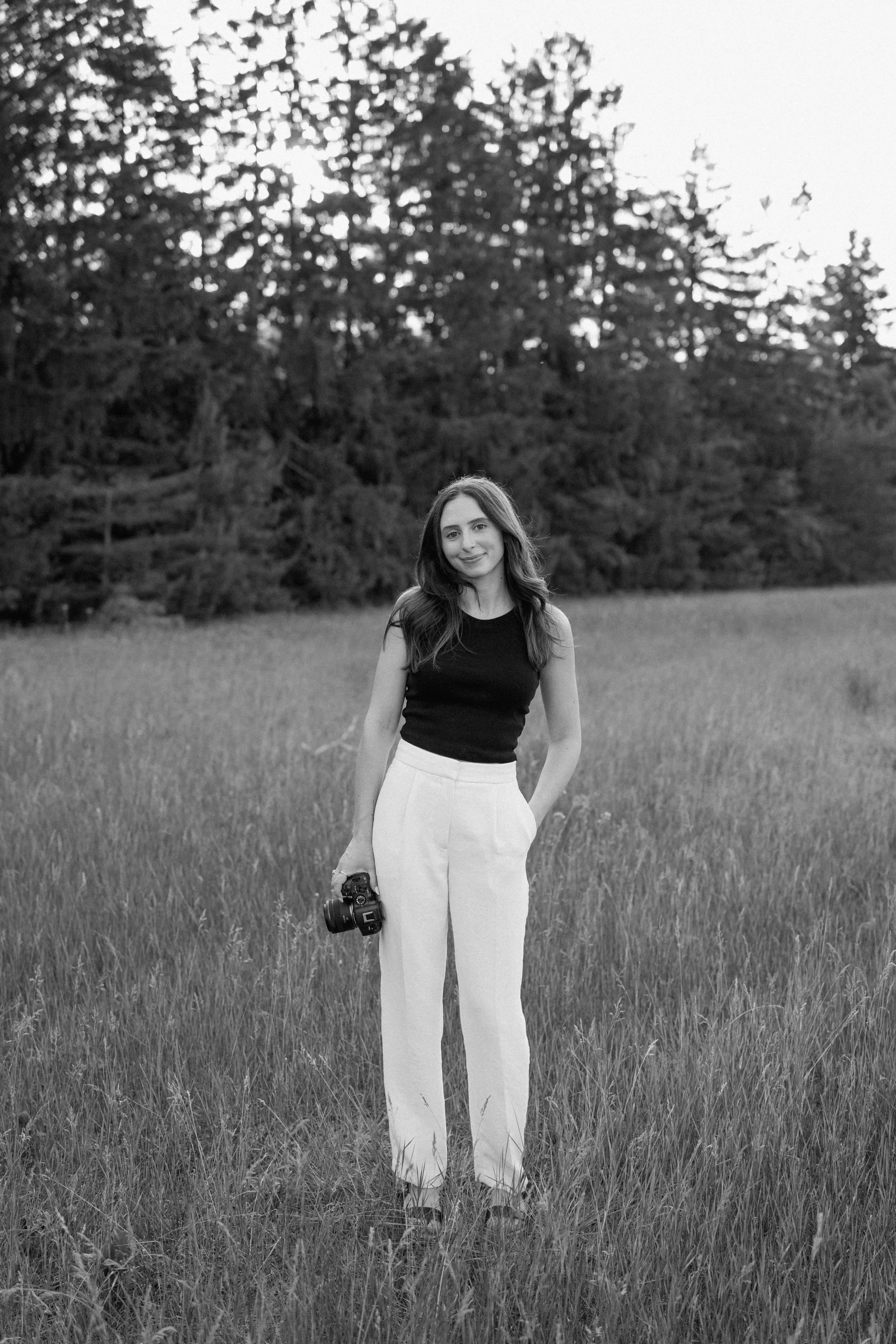 A woman standing in a grassy field holding a camera, with trees in the background, in black and white.