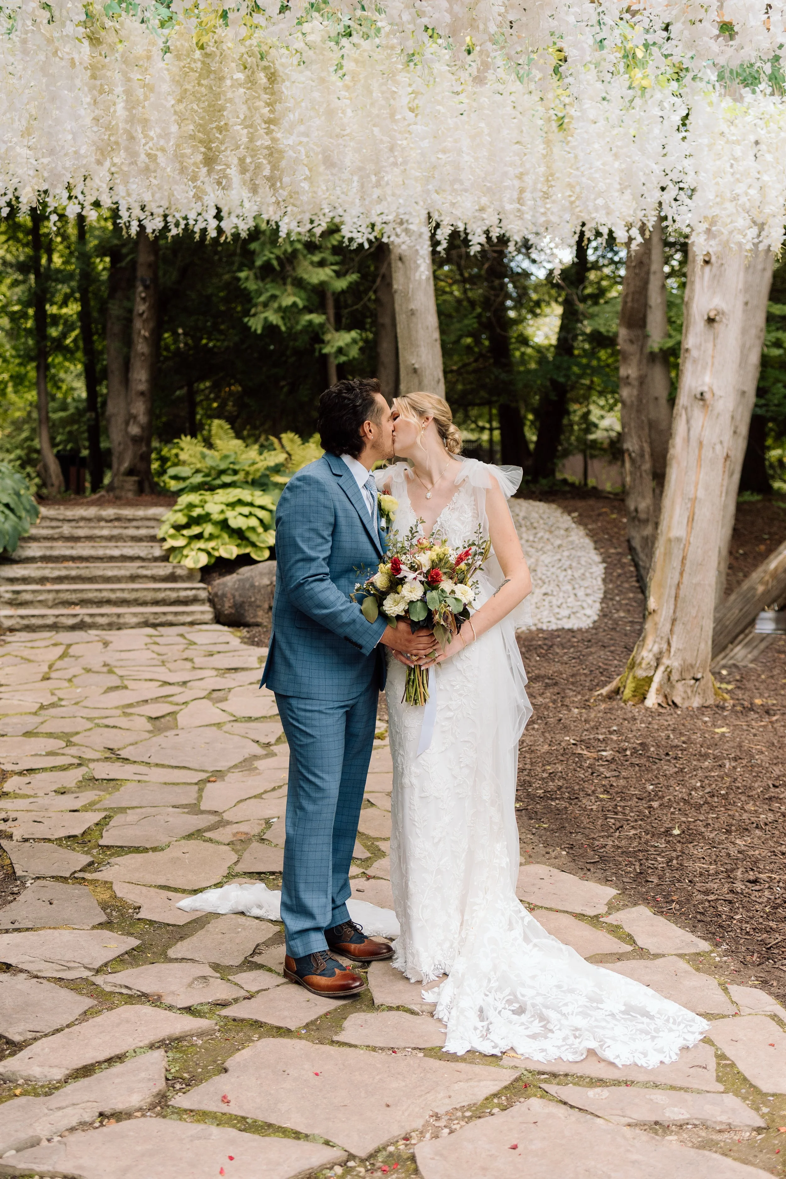 A bride and groom sharing a kiss outdoors during their wedding ceremony under a floral arch with white hanging flowers, surrounded by trees and a stone pathway.