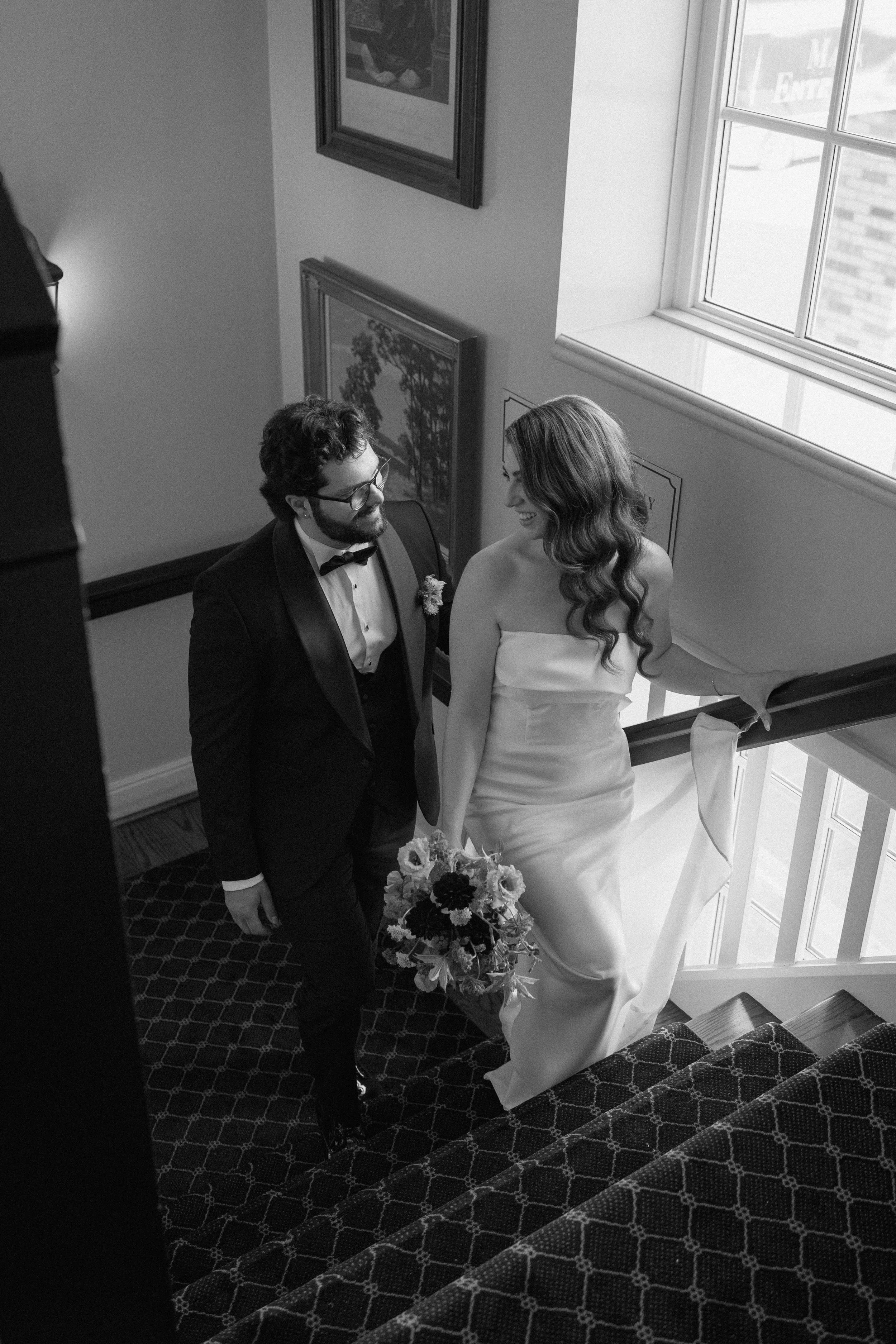 A black and white photograph of a bride and groom ascending a staircase, looking at each other and smiling. The bride is holding a bouquet of flowers, and the groom is wearing glasses and a tuxedo with a bow tie. Sunlight streams in through a window 