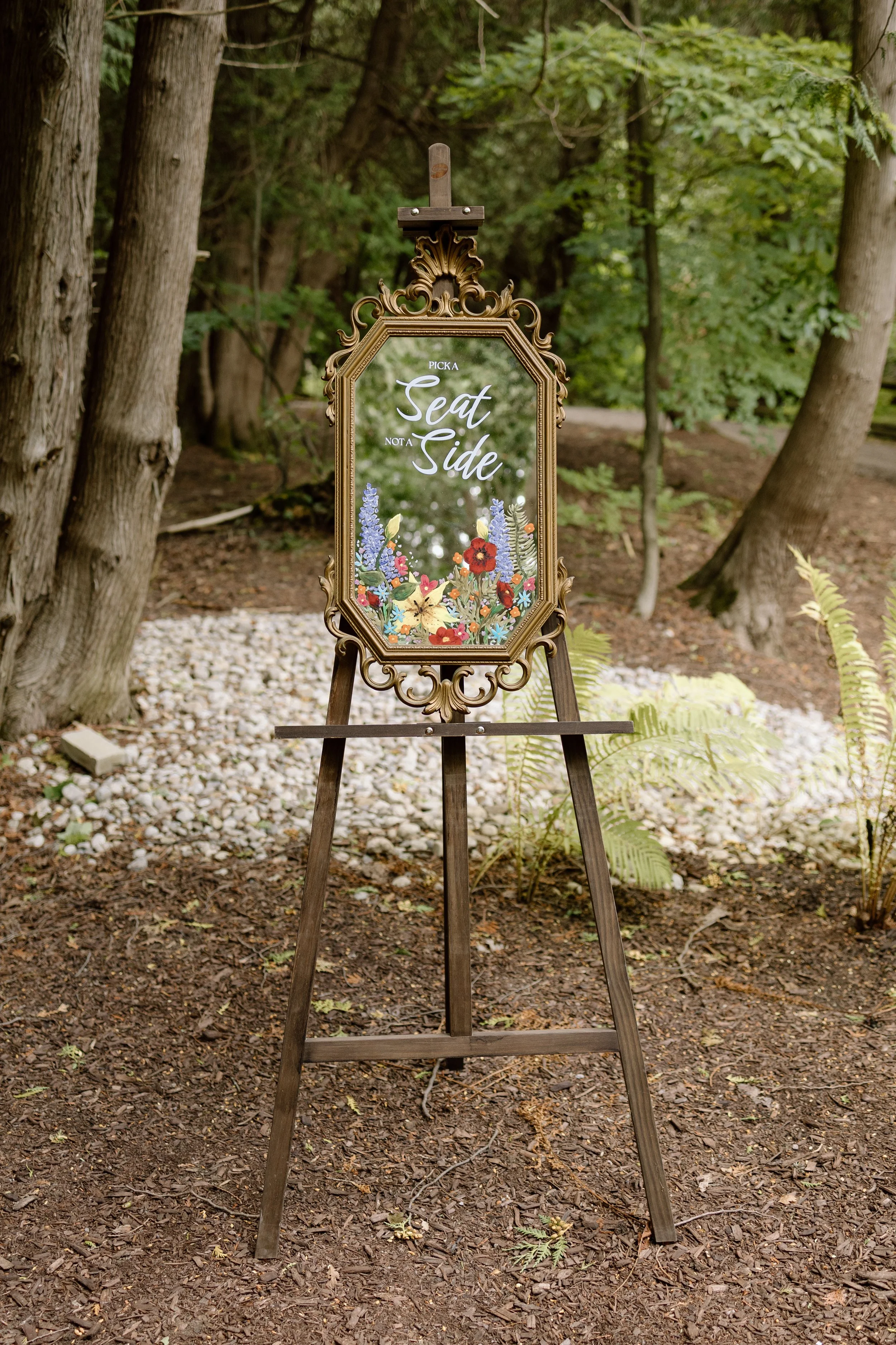 A decorative gold-framed mirror sign on a wooden easel displays the message 'Picka Seat Not a Side' with floral illustrations at the bottom, set outdoors among trees and foliage.