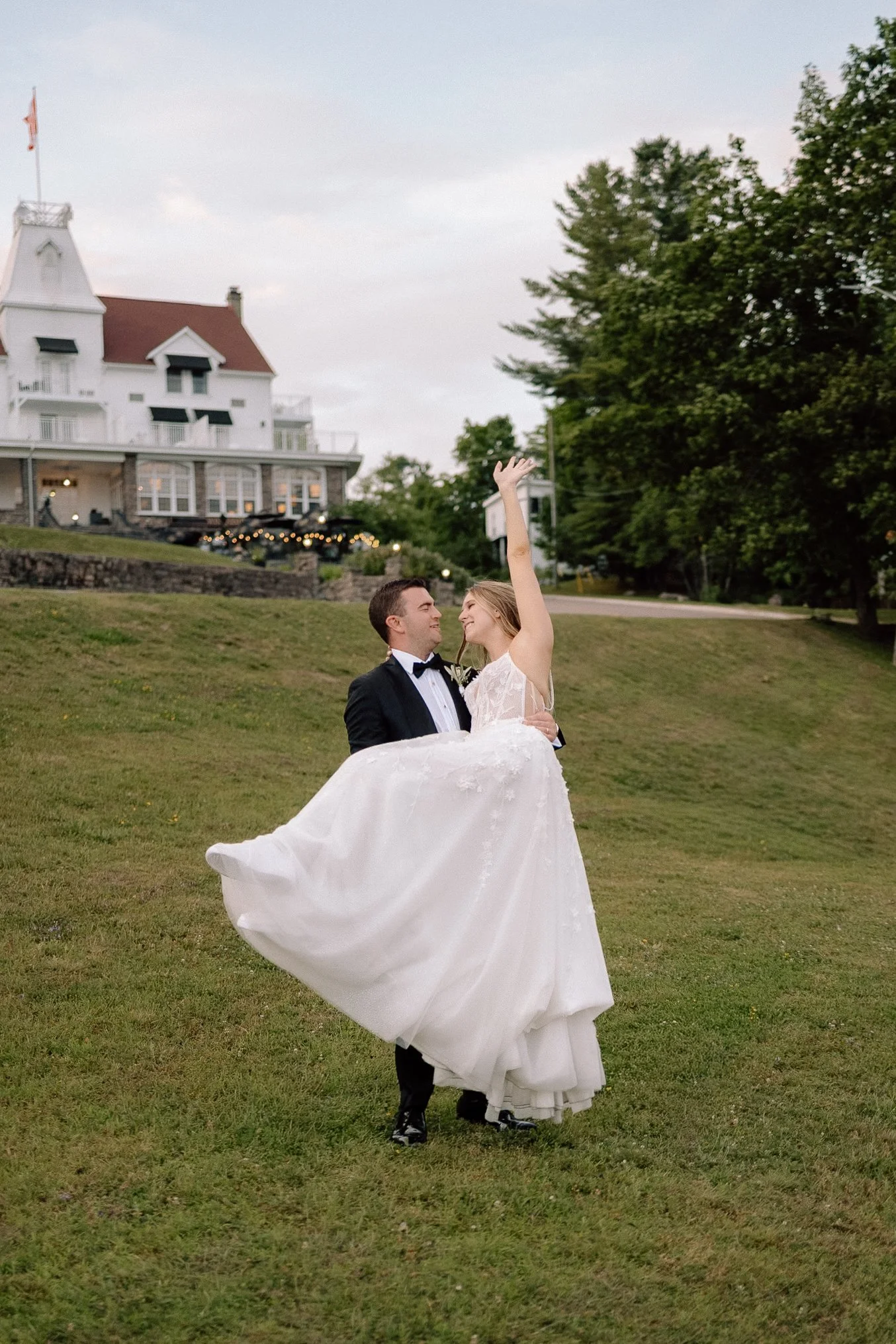 A bride in a white wedding dress being lifted and spun by a groom in a black tuxedo on a grassy lawn with a large white house and trees in the background during an outdoor wedding.