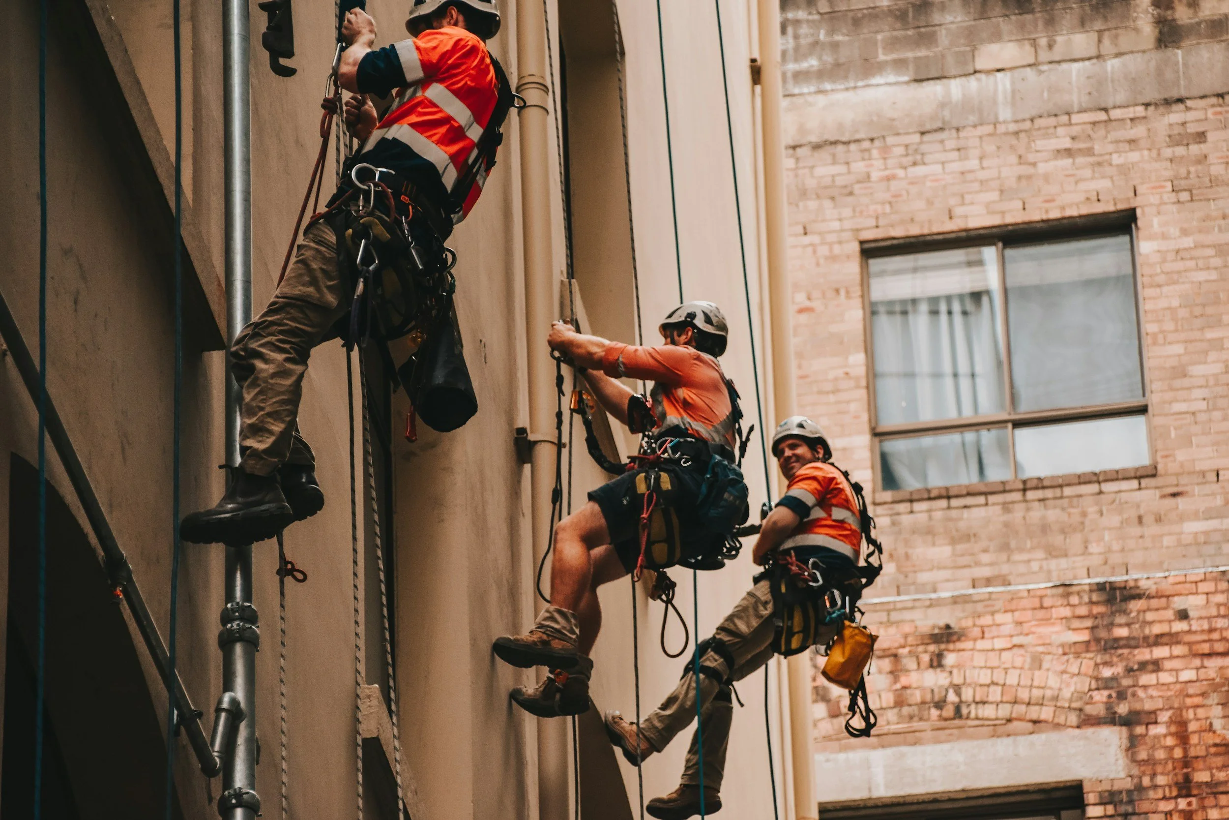 Three rescue workers wearing helmets, safety vests, and harnesses are rappelling down a building exterior to perform a rescue operation.