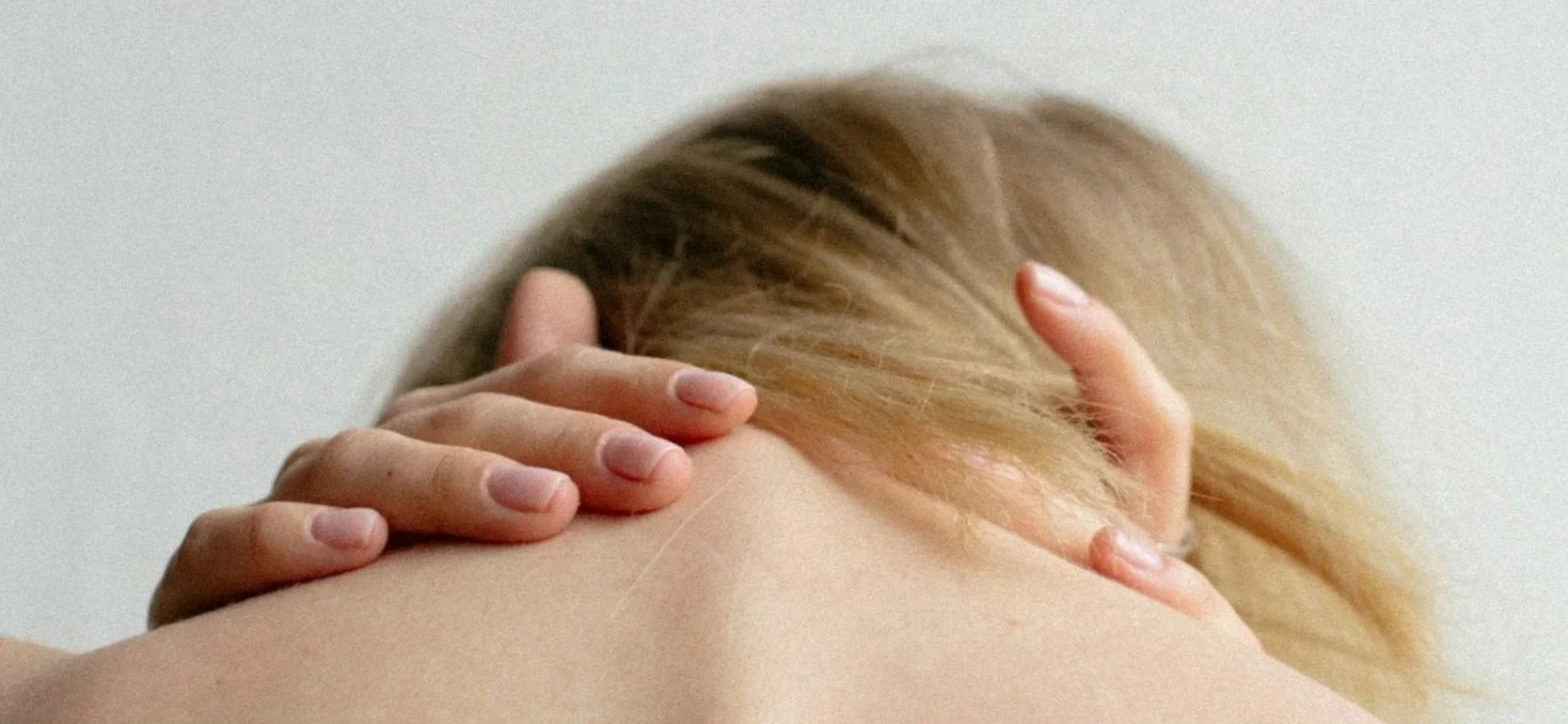 A person with long blonde hair touching their neck and shoulder, viewed from behind, against a plain light background.