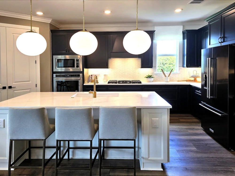 Modern kitchen with white island, three beige chairs, black cabinets, stainless steel appliances, and pendant lighting.