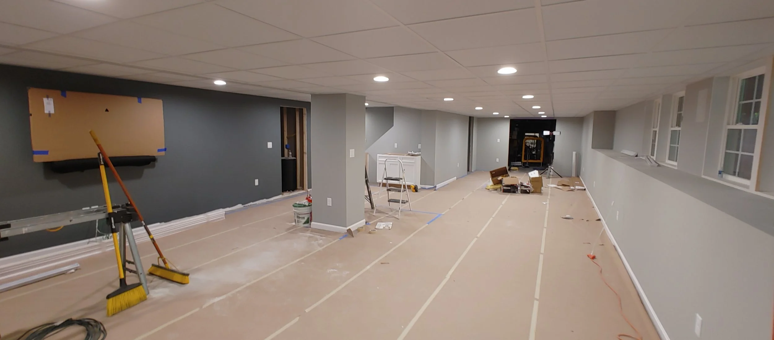 basement renovation with construction tools, cardboard, and ladders, walls painted gray, ceiling with recessed lighting, and multiple windows on the right side.