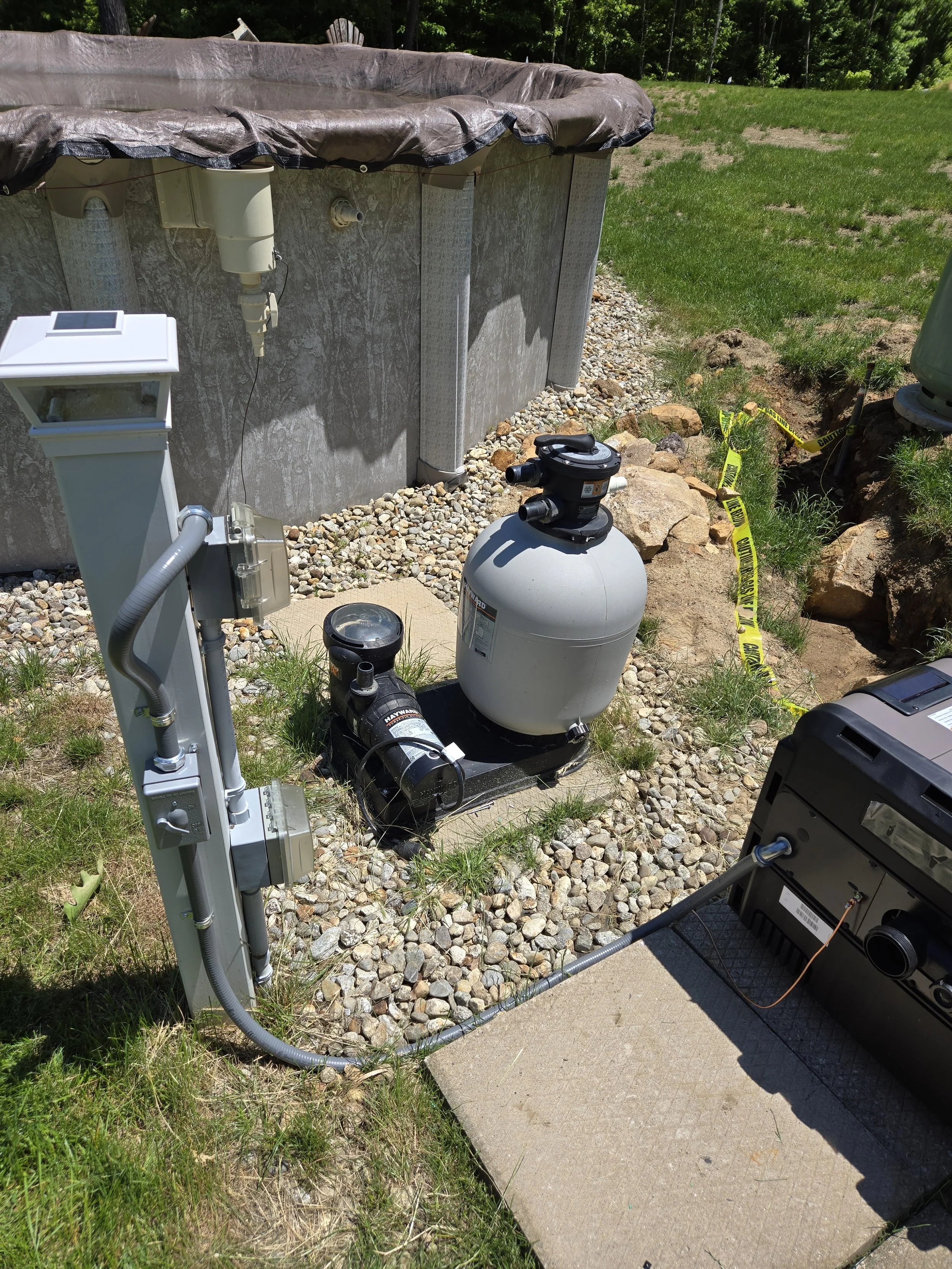 An in-ground swimming pool filtration system with pump, sand filter, and control panel outside near a concrete patio, with rocks and grass around. pool heater install.