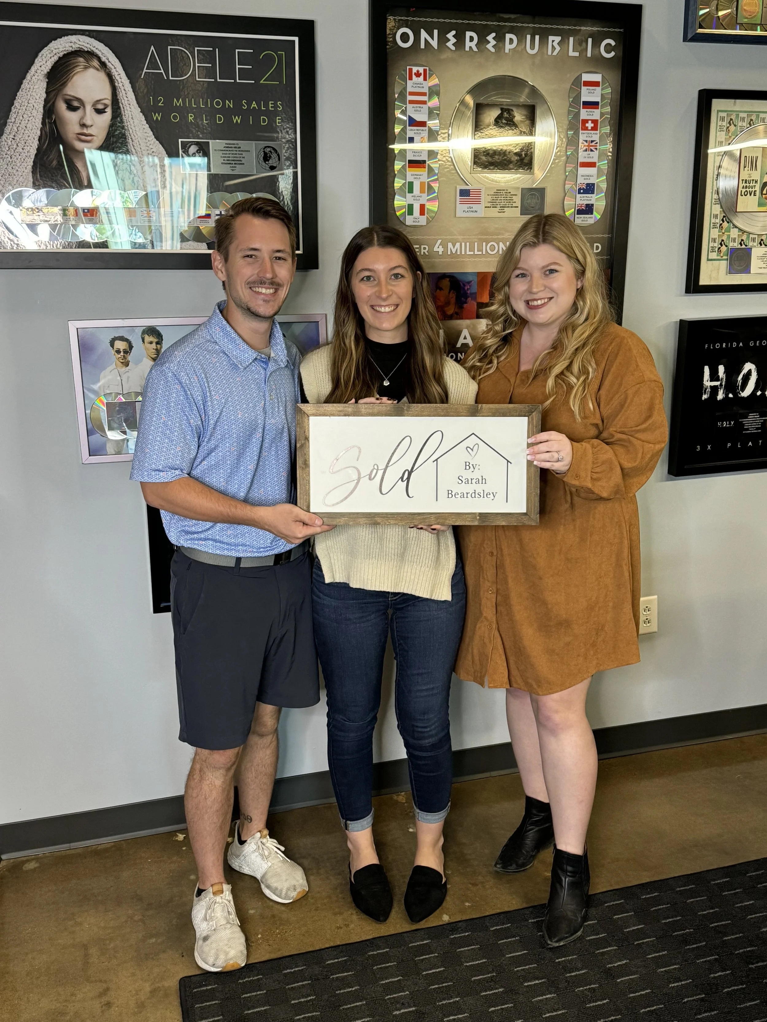 Three people standing indoors, holding a framed sign that says 'Sold' with a name 'Sarah Beardsley.' They are smiling, and there are framed music awards and posters on the wall behind them.