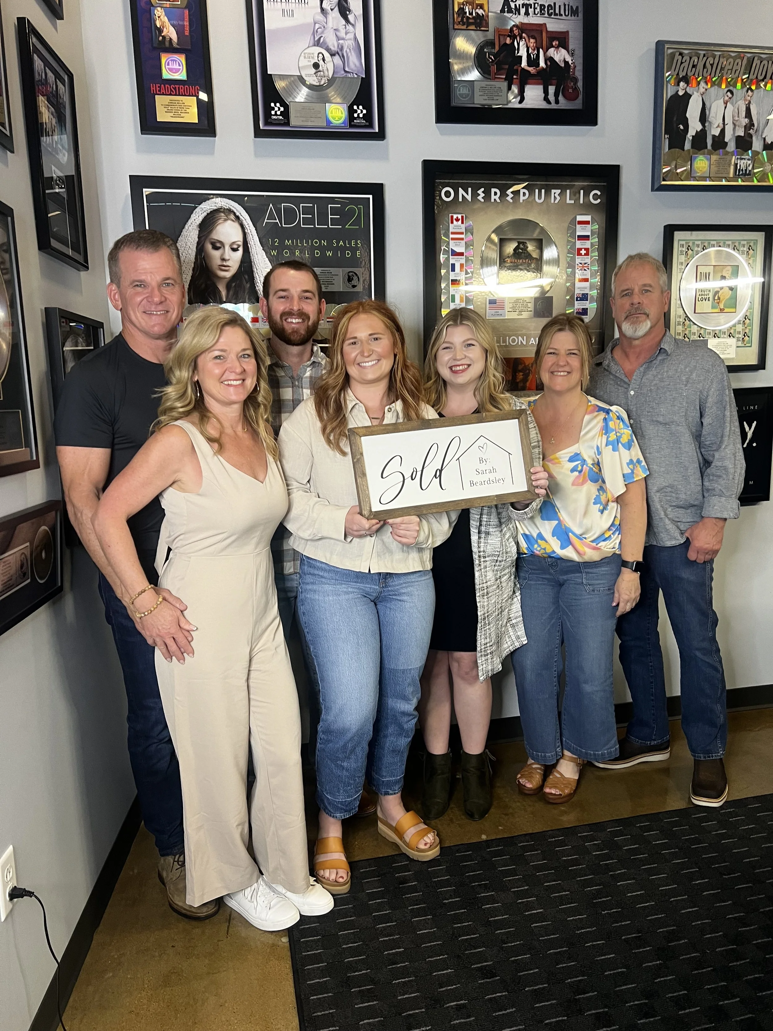 Group of seven people celebrating a sold house, holding a sign that says "Sold by Sarah Beardsley," in a room with music awards and framed posters on the wall behind them.