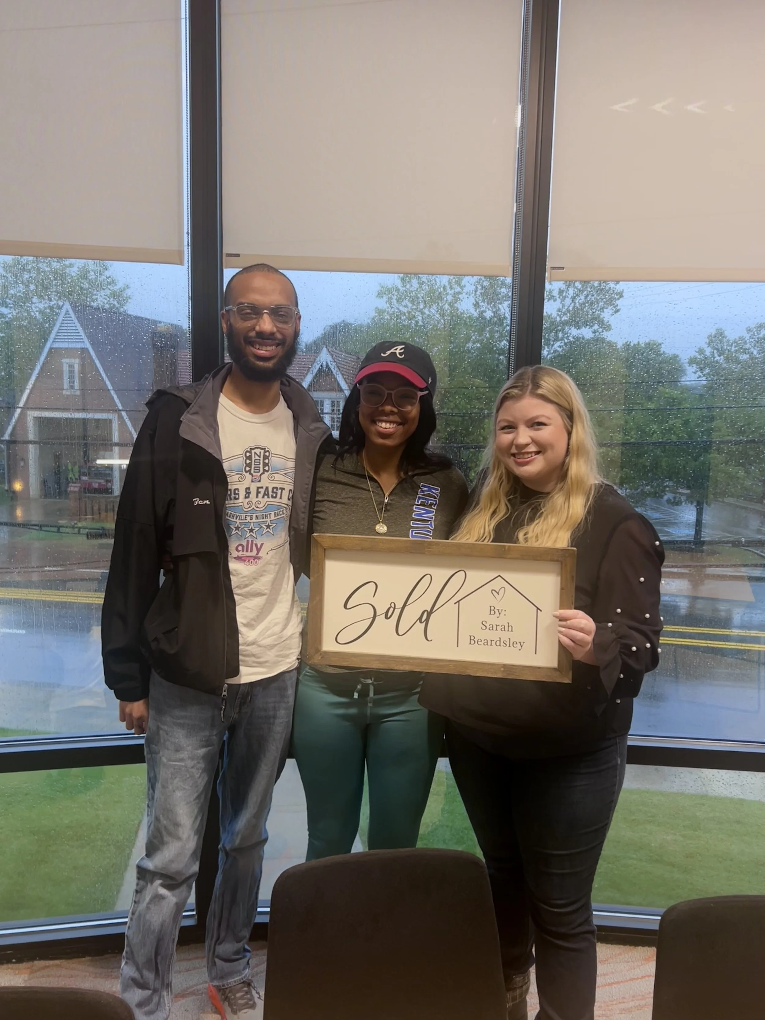 Three people standing inside by large window, holding a wooden framed sign that says 'Sold' with the seller's name, Sarah Beardsley, written on it. Outside rainy day with houses and trees visible.