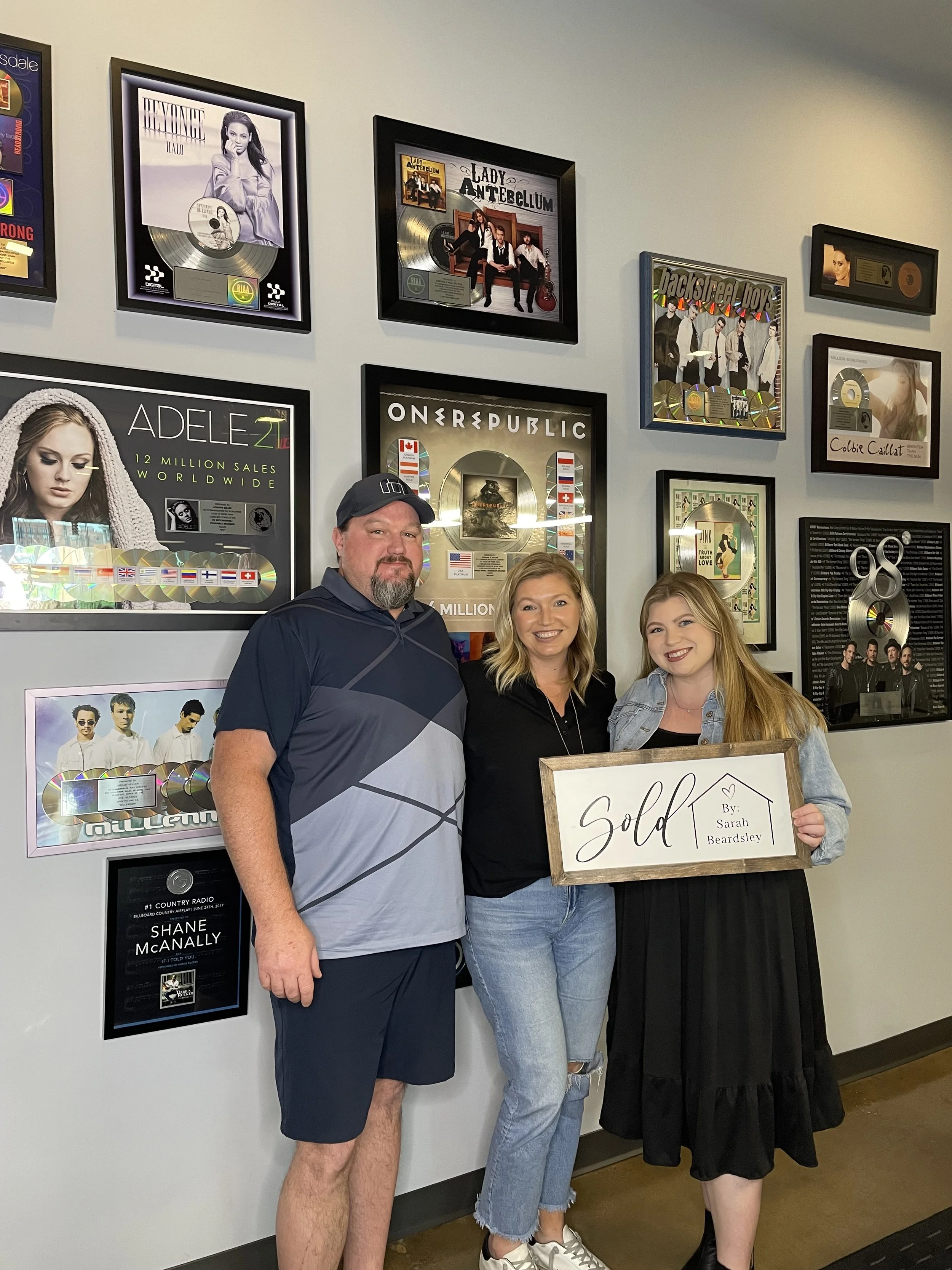Three people standing together in a hallway with framed music awards and posters on the wall. The woman on the right is holding a wooden sign that reads "Sold" with a small heart and the name Sarah Beardsley. All three are smiling.