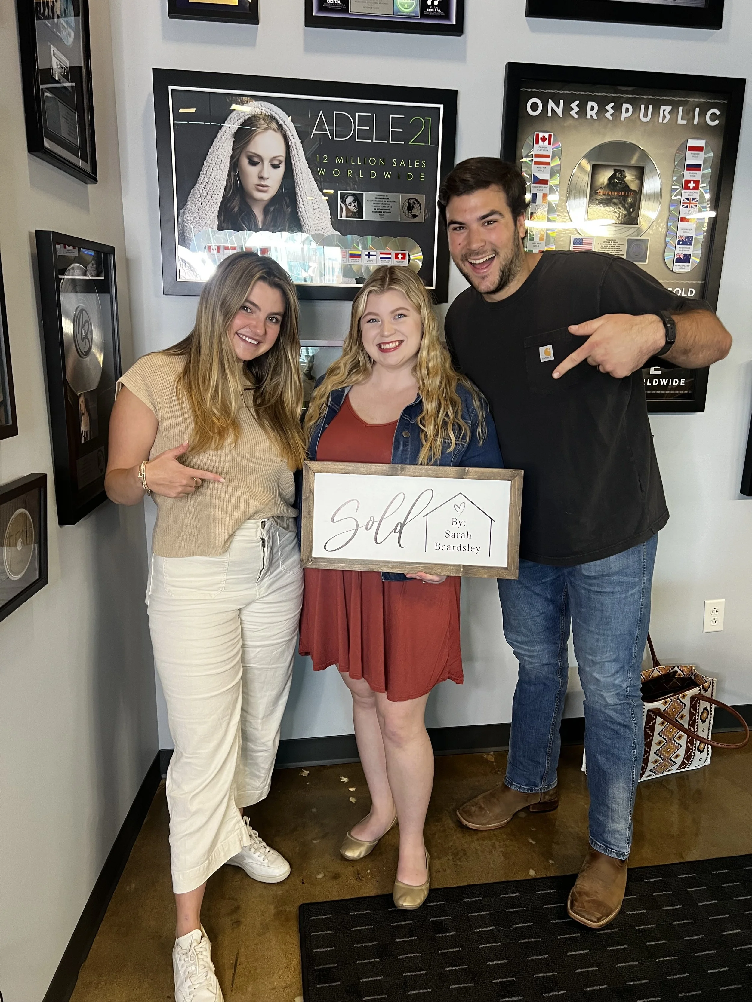 Three people standing indoors, holding a sign that says 'Sold by Sarah Beardsley'. They are smiling and pointing to the sign. The woman in the middle has blonde hair and is wearing a red dress and gold shoes. The woman on the left has long blonde hai