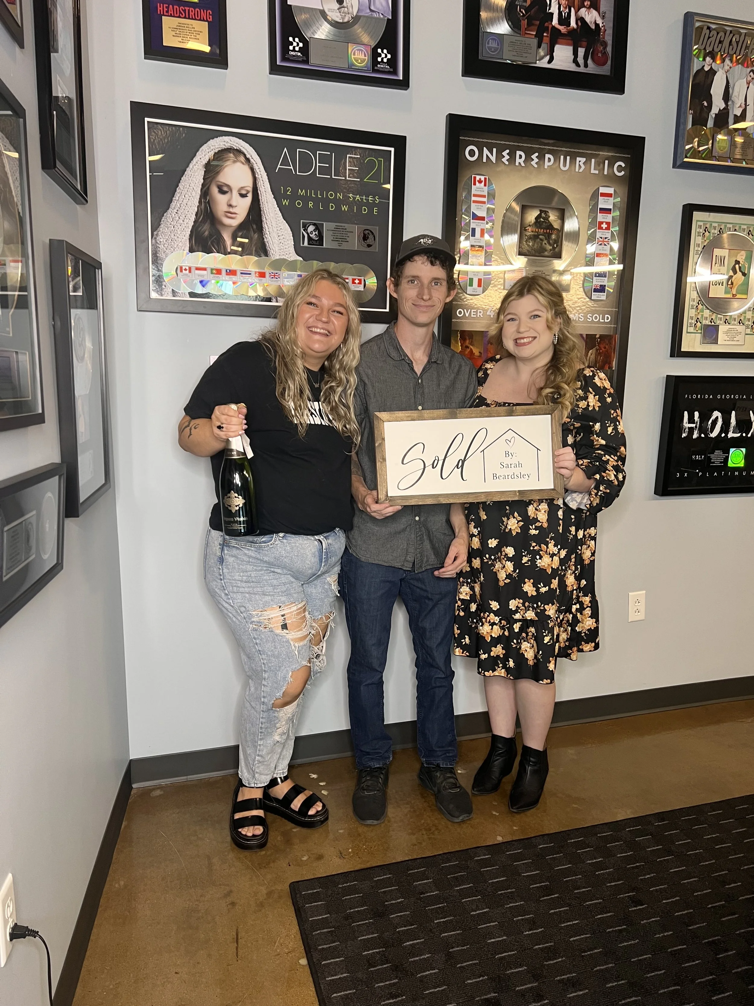 Three people standing indoors, holding a framed sign that reads "Sold by Sarah Beardsley." The person in the middle appears to have just sold a house. The woman on the left is holding a bottle of champagne, and the woman on the right is wearing a flo