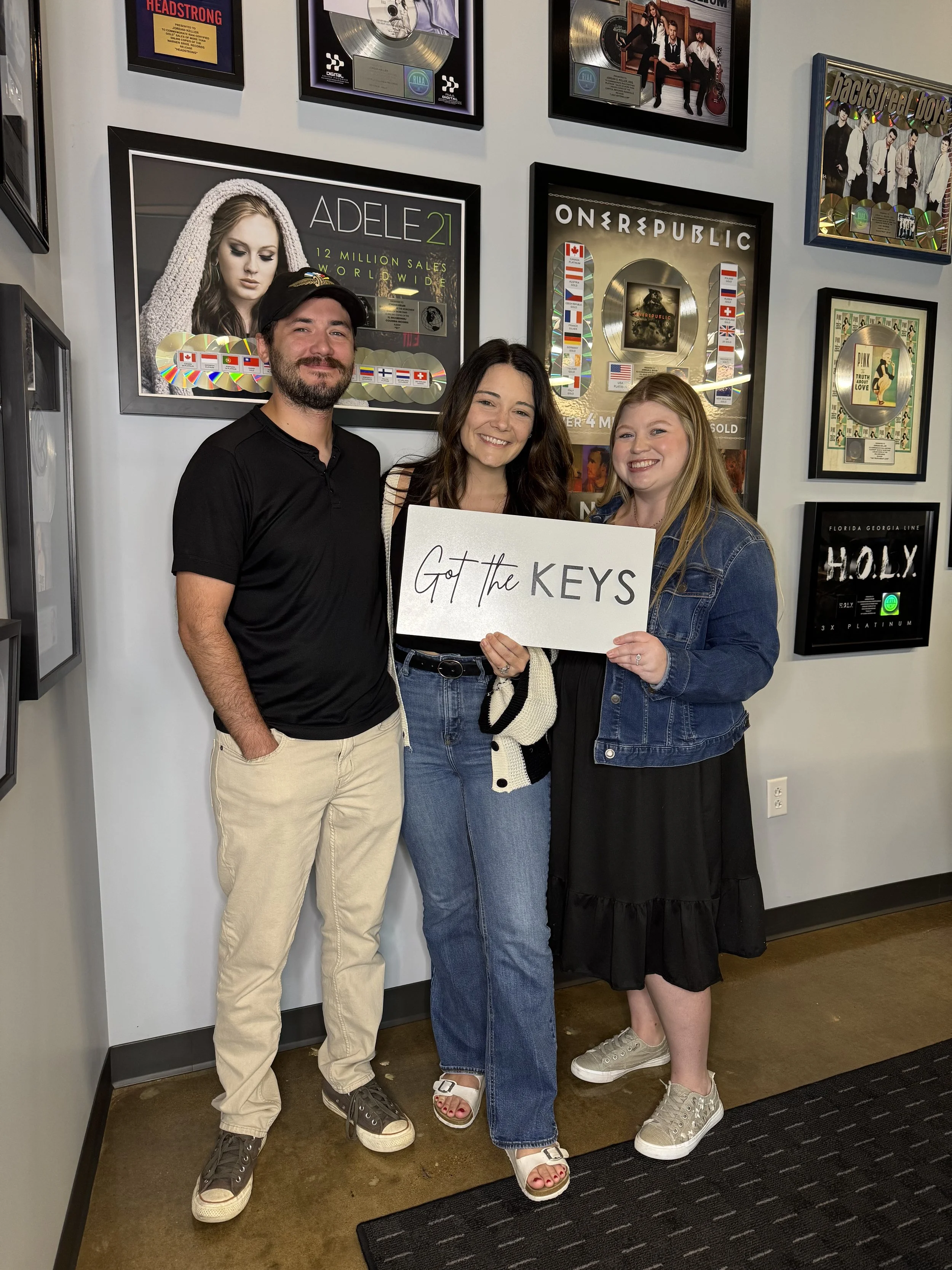 Three people standing together in a room decorated with music awards and records on the wall. They are smiling, with the woman in the middle holding a sign that says 'Got the KEYS'. The man on the left has a beard, and all three are casually dressed.