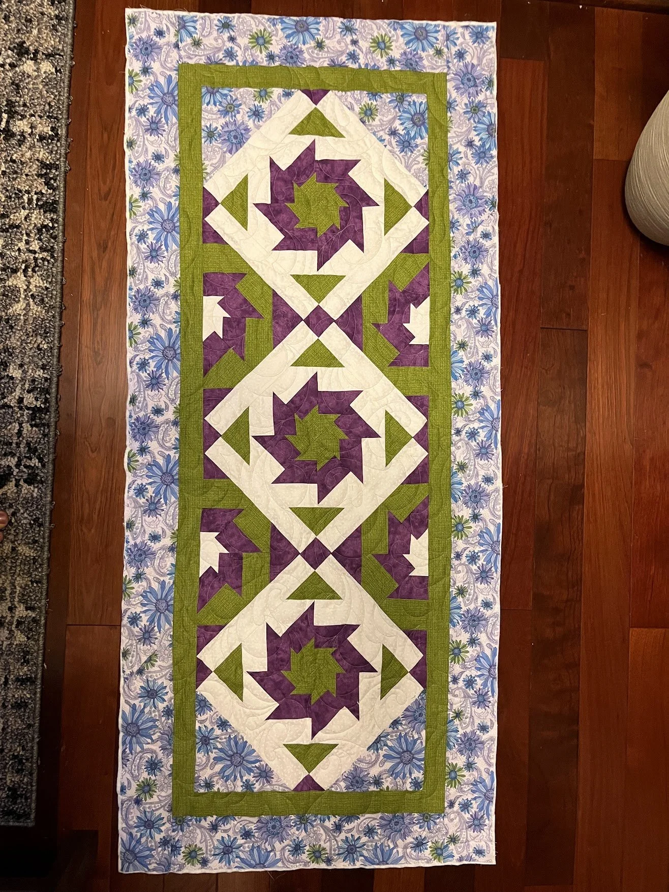 Colorful quilt with purple, green, and white geometric patterns on a floral border, laid out on a wooden floor.
