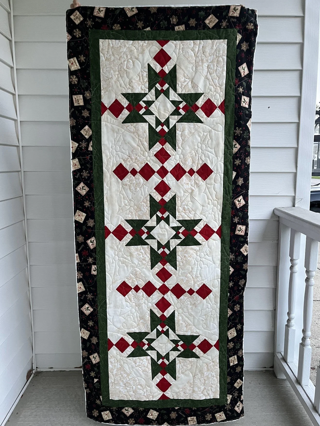 A vertical rectangular quilt with a Christmas theme featuring a pattern of stars and diamonds in red, green, and white fabrics, bordered with black fabric decorated with small images of Christmas ornaments, all hanging on a porch with white siding an