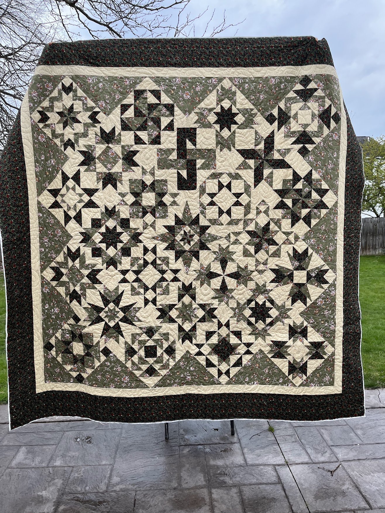 A patchwork quilt with a complex star pattern, featuring a mix of dark and light fabric pieces, bordered by dark fabric with small floral prints, hanging outdoors on a stand against a background of blue sky and trees.