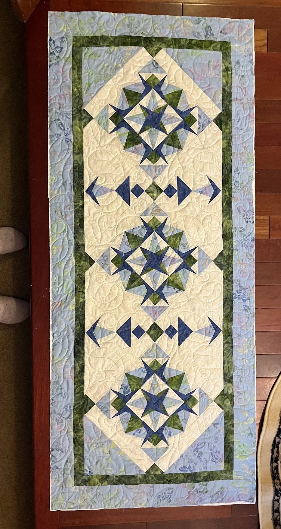 A long rectangular quilt with a geometric pattern, featuring blue, green, and cream colors, laid on a red wooden surface.