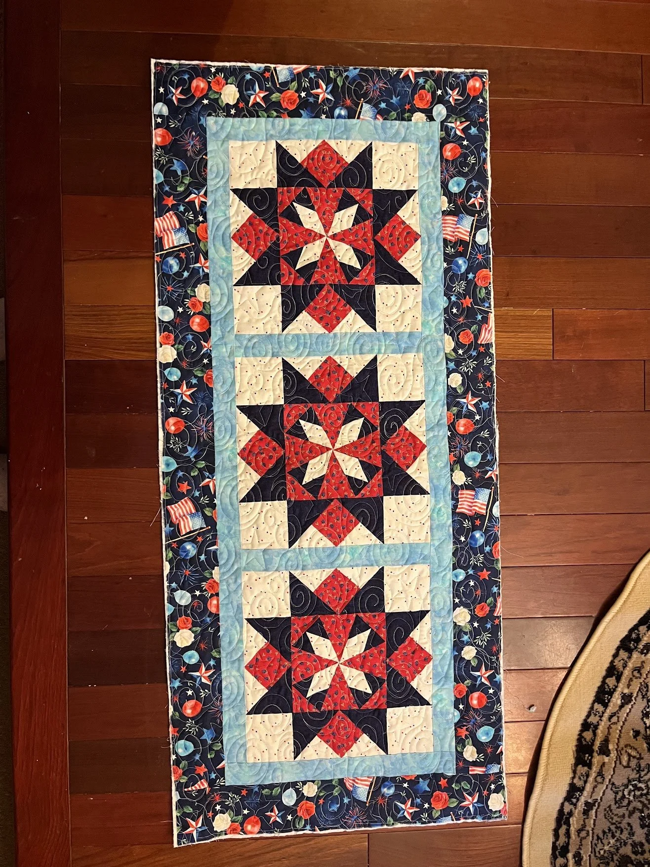 Patriotic quilt with star and geometric patterns in red, white, blue, and black, featuring a border with fireworks and American flags on a wooden floor.
