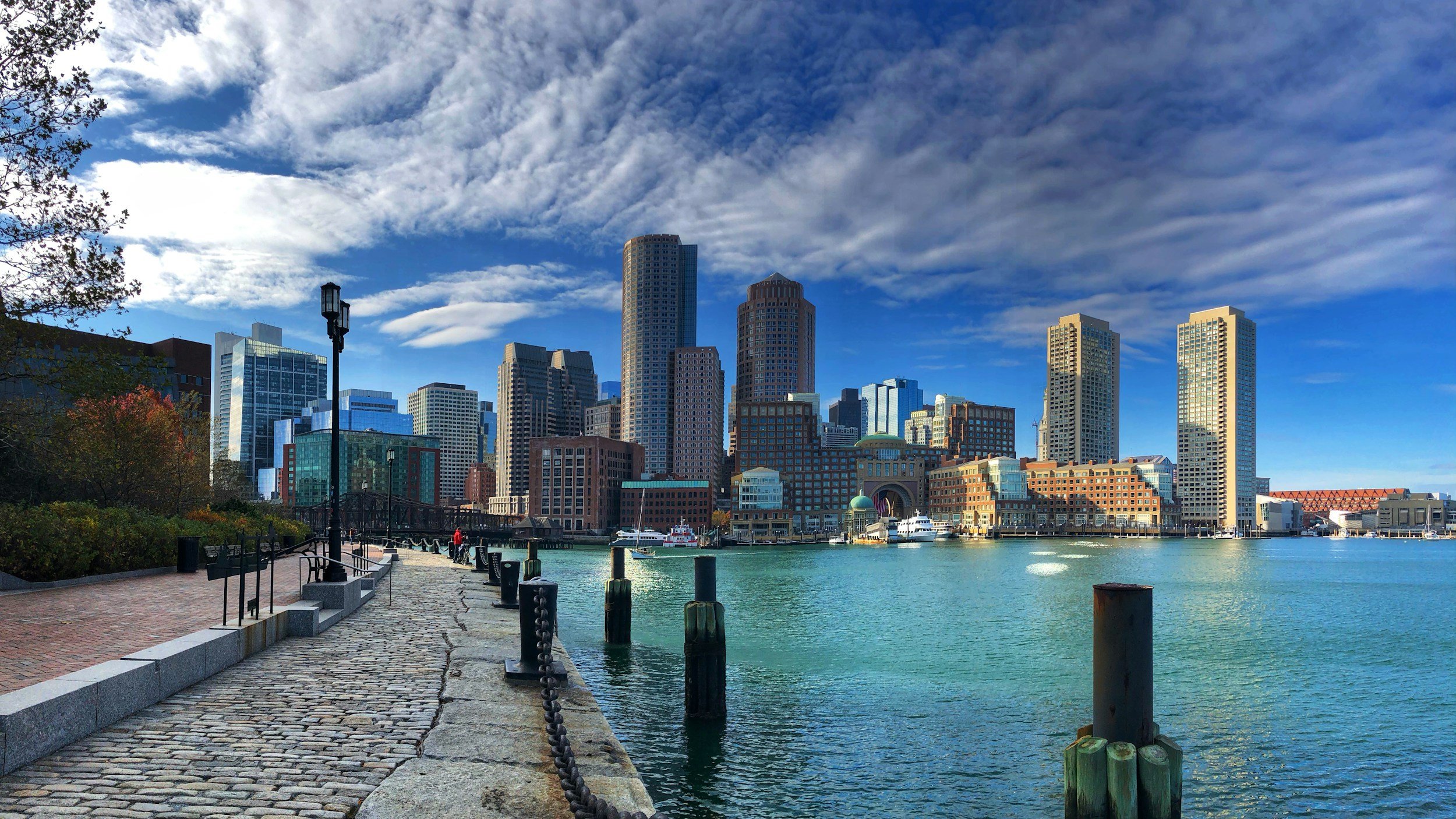 City skyline with tall skyscrapers viewed across a harbor, with a brick and stone walkway along the water and cloudy blue sky overhead.