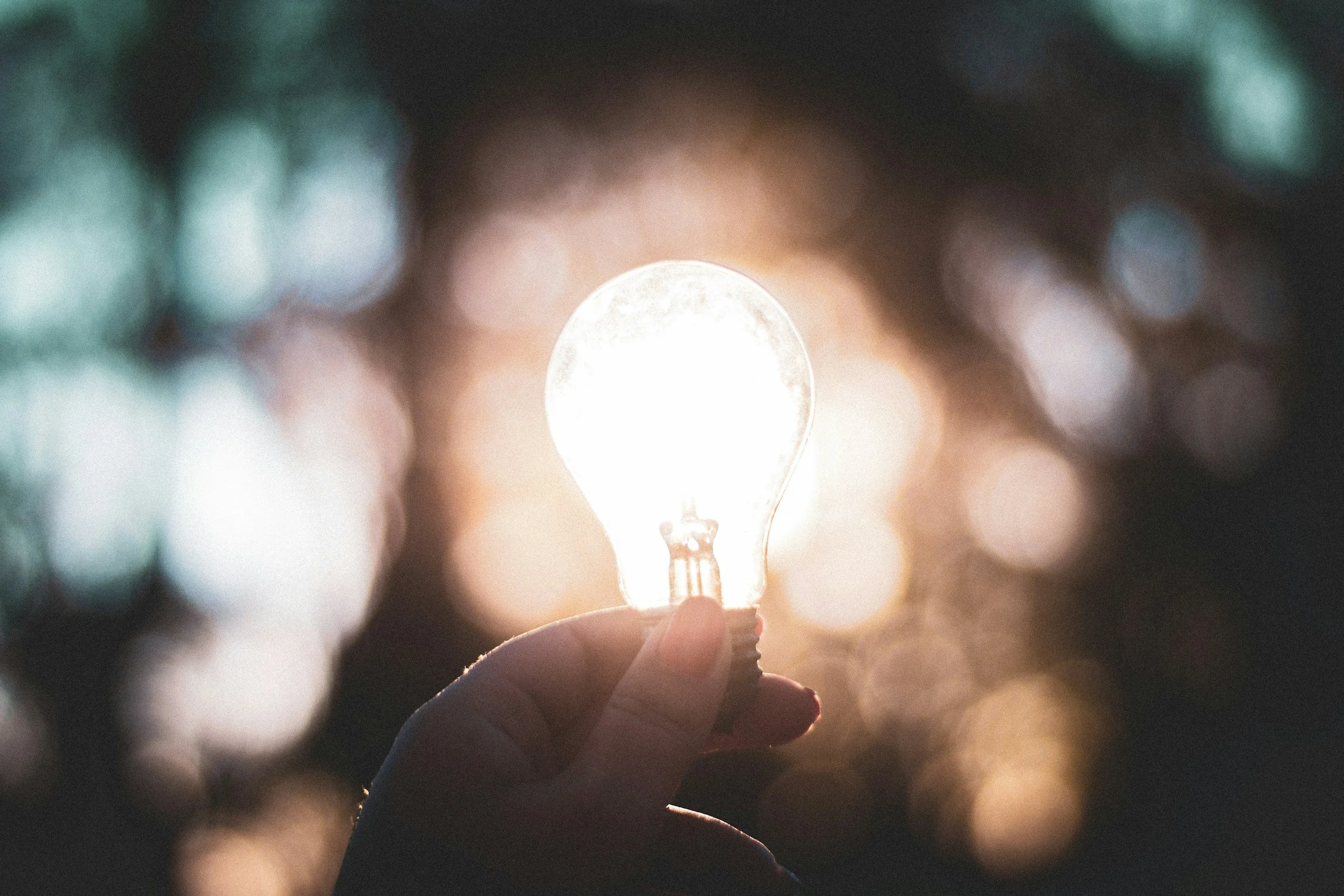 A hand holding a light bulb in front of blurred background lights.