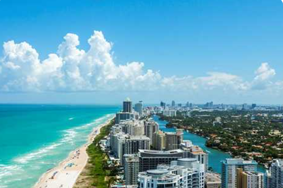 Aerial view of a coastal city with high-rise buildings along the shoreline, turquoise ocean, sandy beach, and partly cloudy sky.