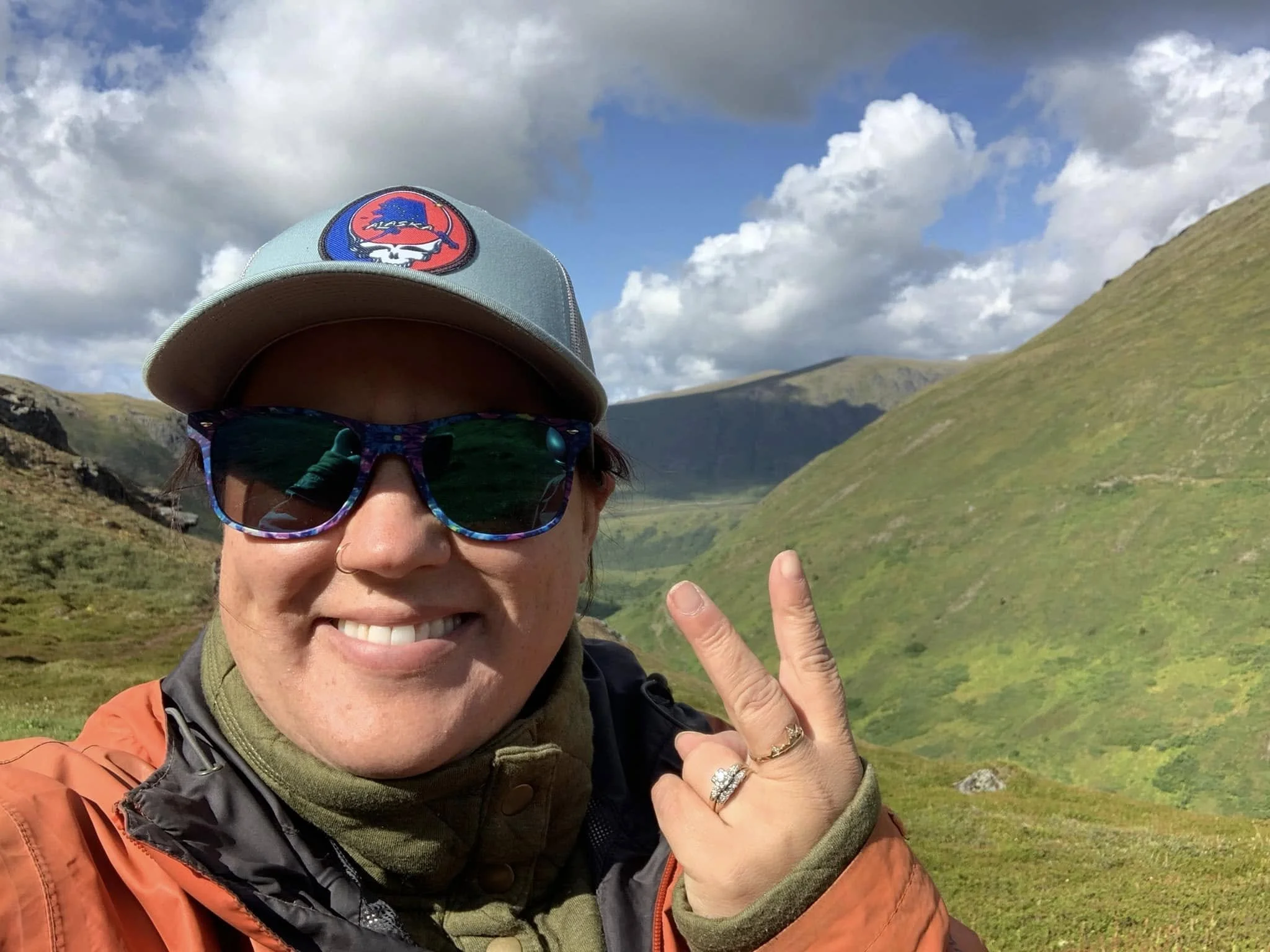 A woman smiling and making a peace sign with her hand, wearing sunglasses, a baseball cap, and outdoor gear, with a background of green hills and a partly cloudy sky.