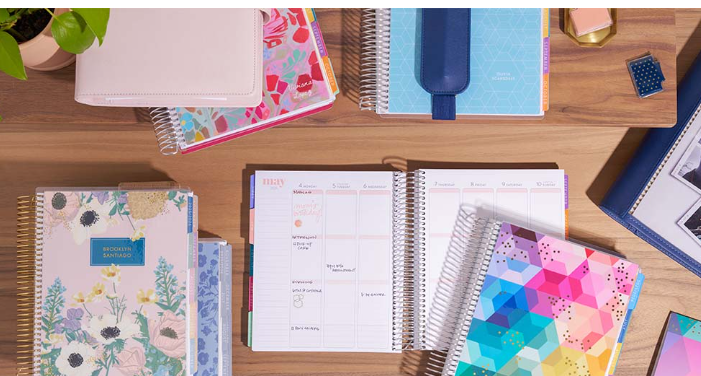 A wooden desk with a variety of colorful planners and notebooks, some open and some stacked, along with a small plant and a gold-colored object.