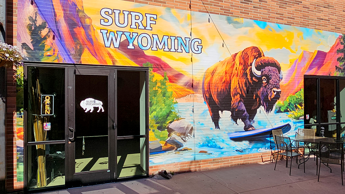Colorful mural on a brick wall depicting a bison surfing a wave with mountains in the background, and the words "SURF WYOMING" in large letters.