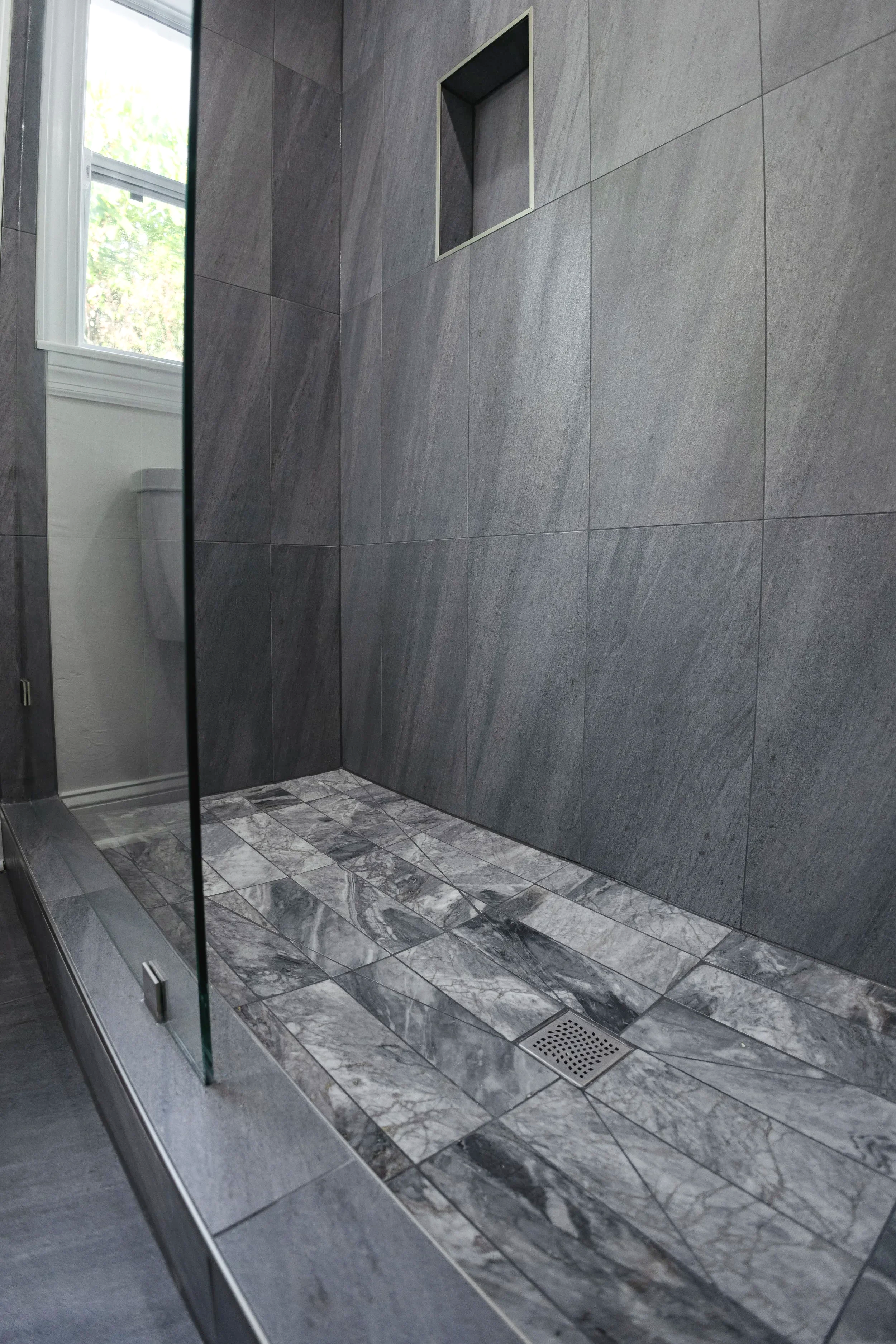 Empty shower with gray tiled walls and marble-patterned floor, glass door, and a small square niche in the wall.
