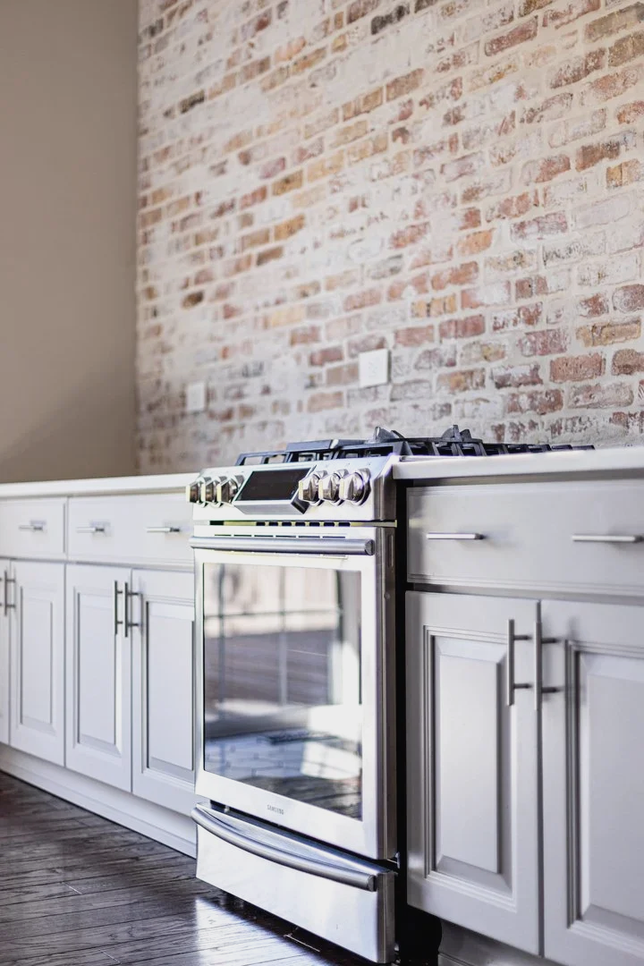 Modern kitchen with white cabinets and a stainless steel stove against an exposed brick wall.