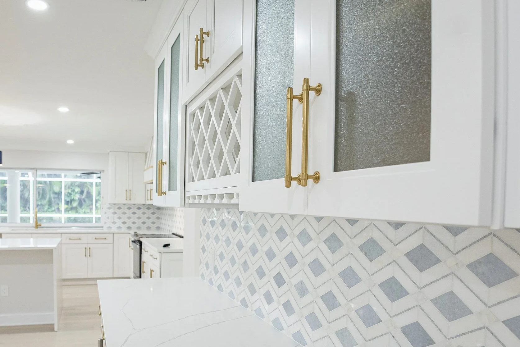 Modern white kitchen with gold handles on cabinets, a geometric patterned backsplash, and a large window letting in natural light.