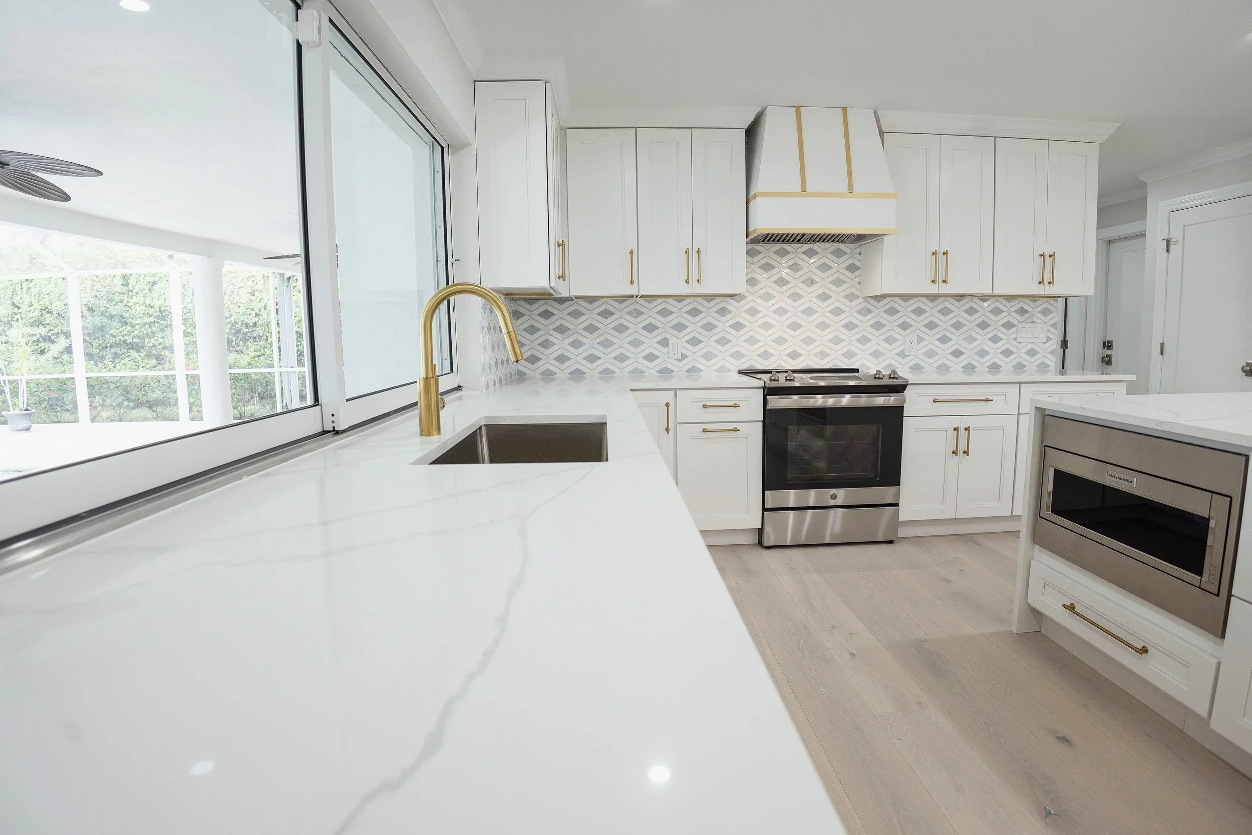 Modern kitchen with white cabinets, marble countertops, stainless steel oven, microwave, and gold accents, overlooking an outdoor patio through large windows.