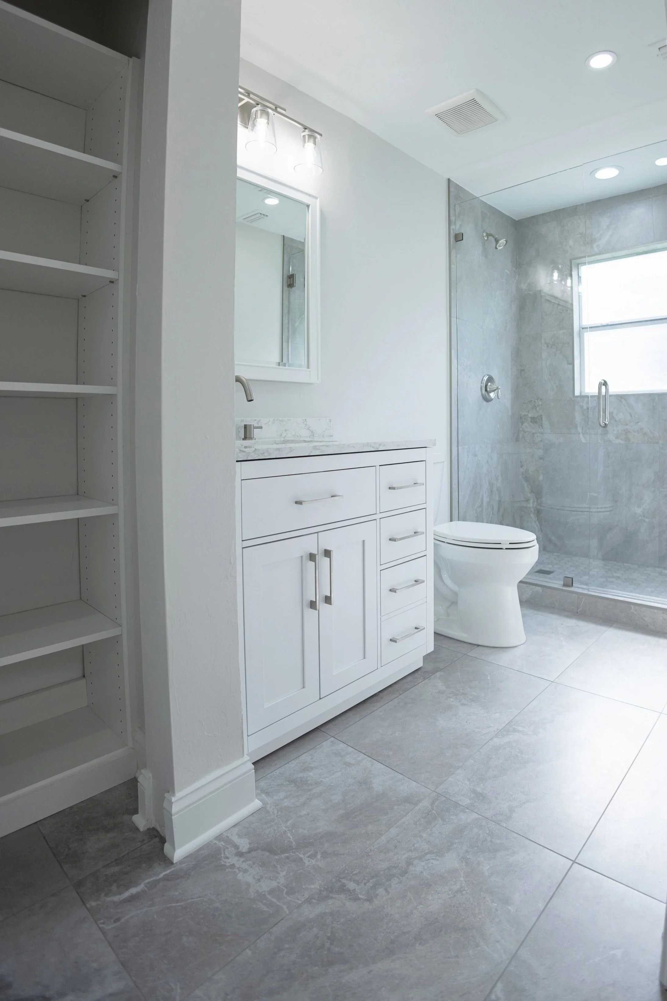 Modern bathroom with white vanity, toilet, tiled shower area with glass door, and large window.