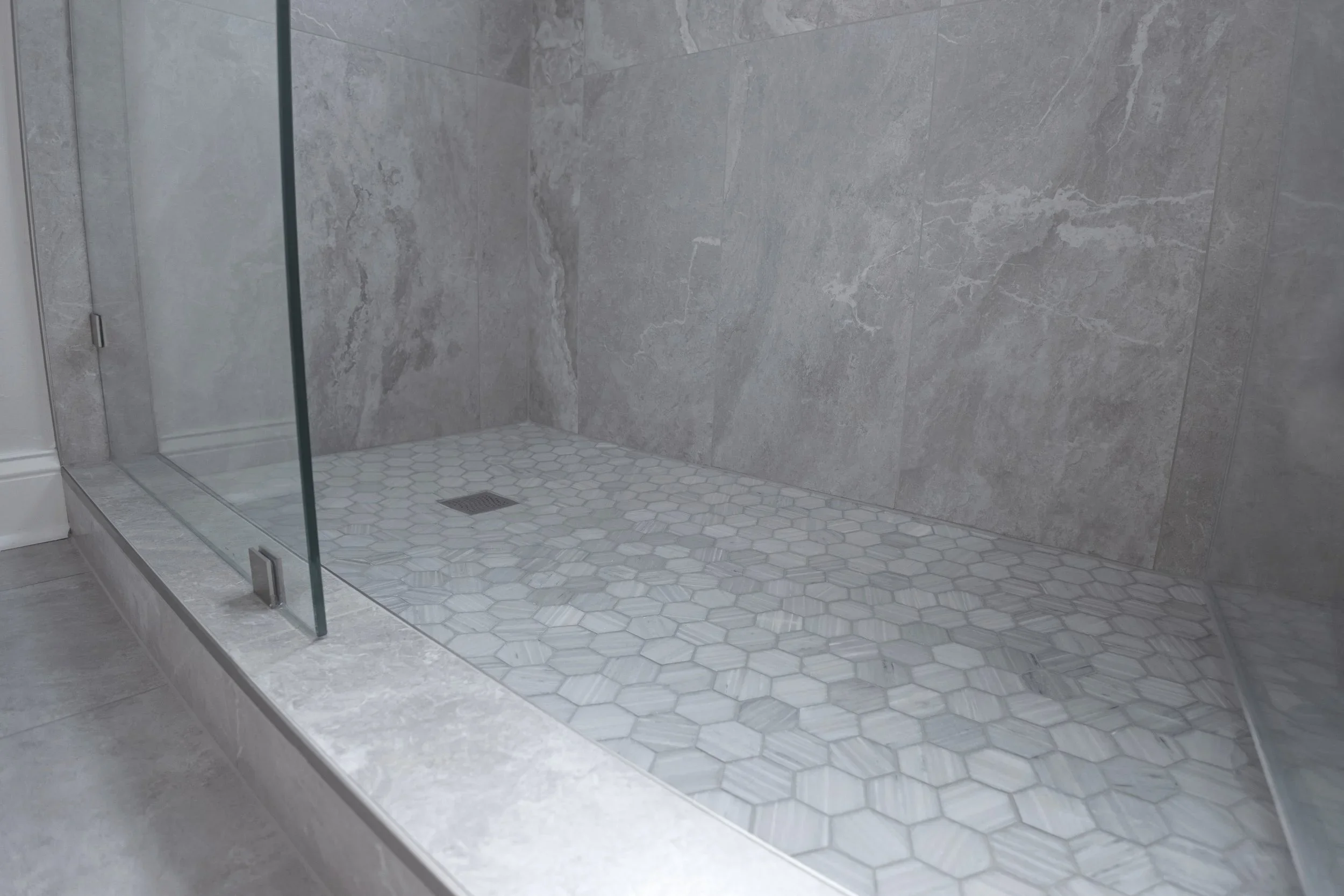 Modern walk-in shower with gray marble walls, white hexagonal floor tiles, and a glass door.