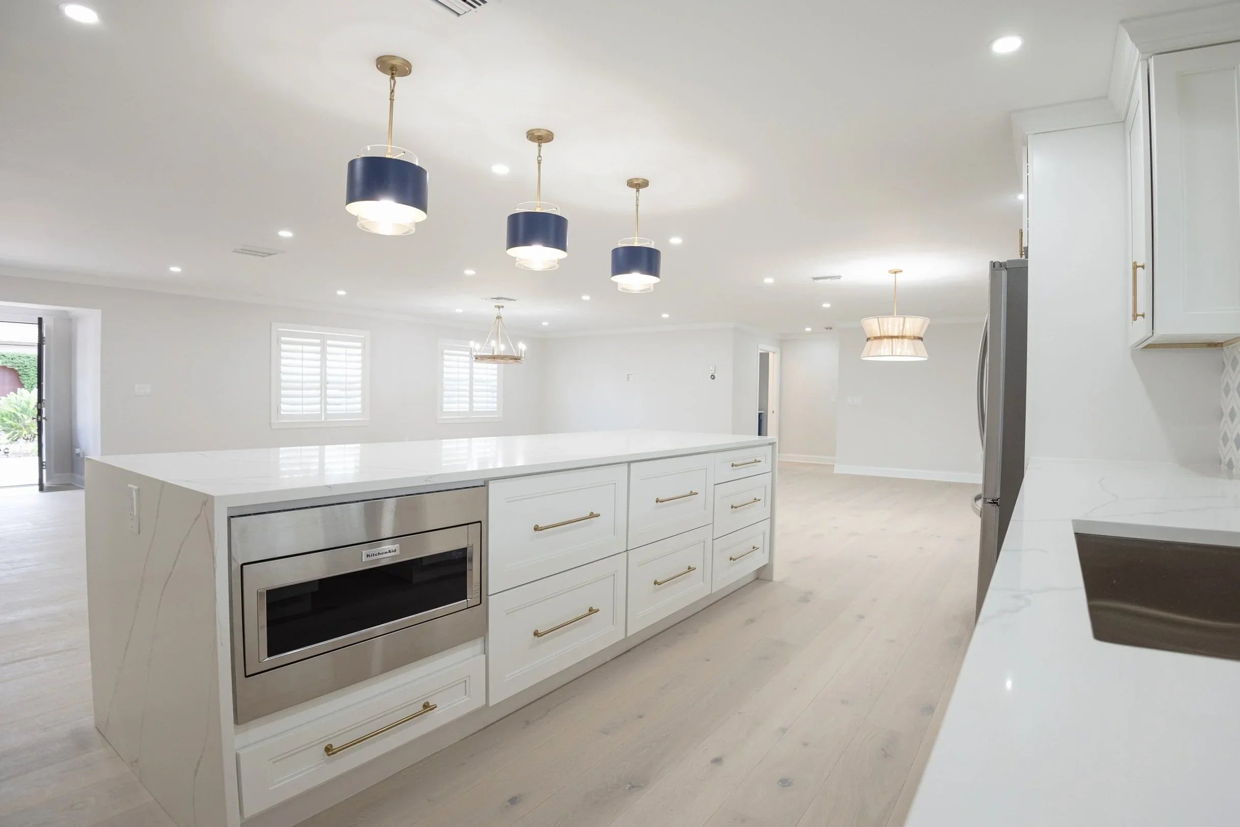 Bright, modern kitchen with white cabinets, marble countertops, stainless steel microwave, gold hardware, and black and gold pendant lights over a large island.