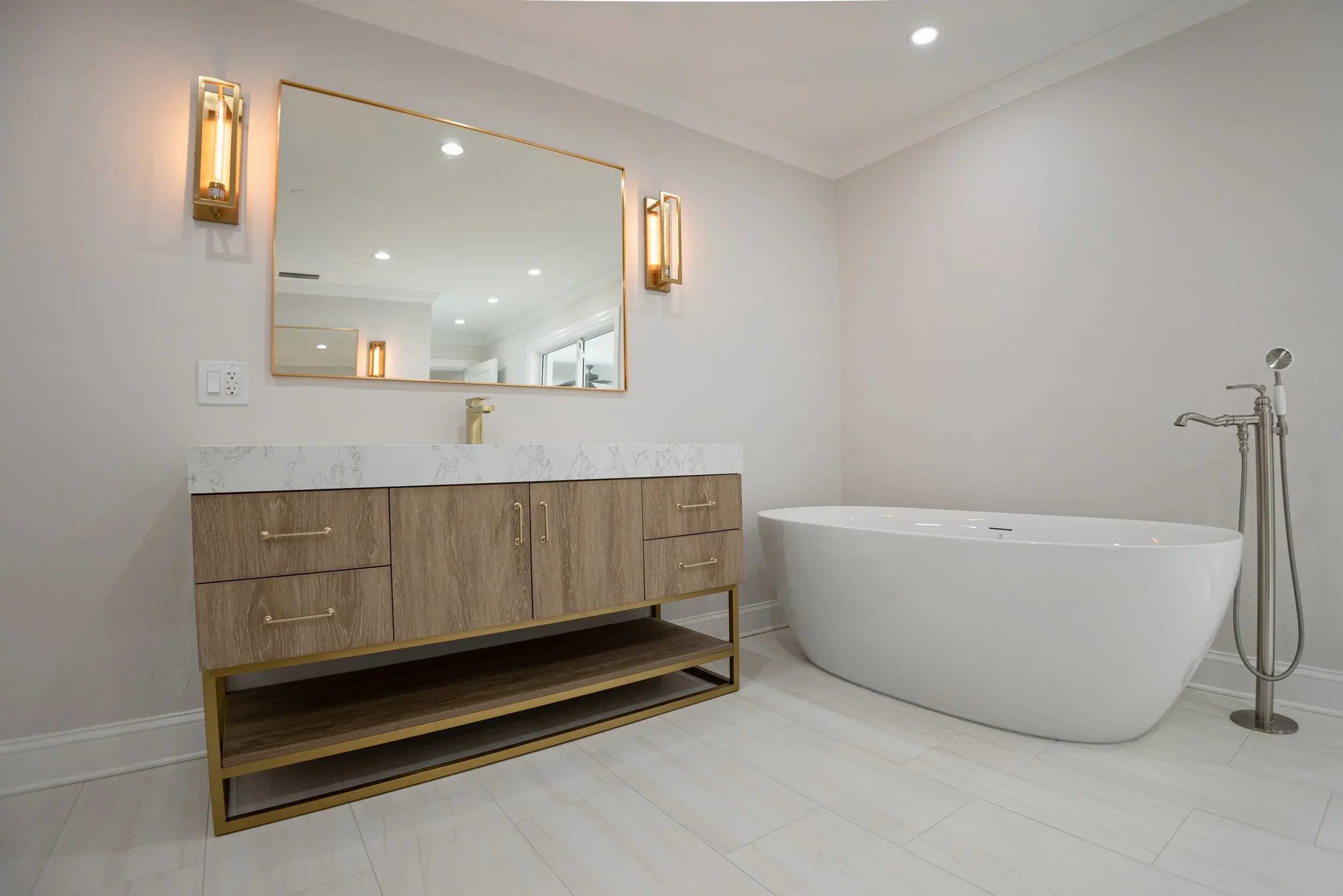 Modern bathroom with white bathtub, wooden vanity with marble countertop, large wall mirror, and wall-mounted gold light fixtures.