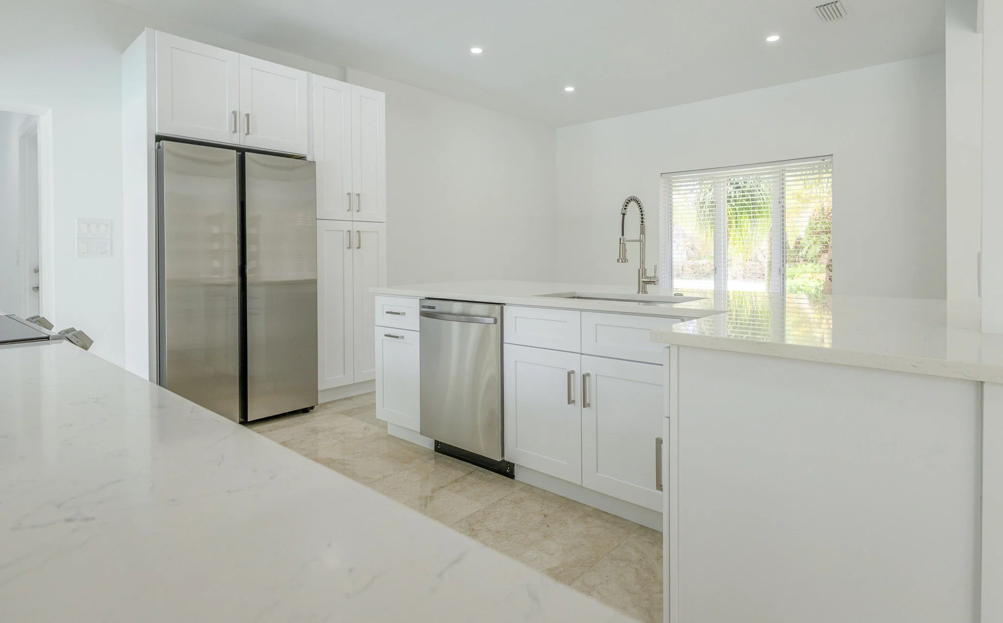 Modern white kitchen with stainless steel appliances, including a double-door refrigerator and a dishwasher, with natural light coming through a window
