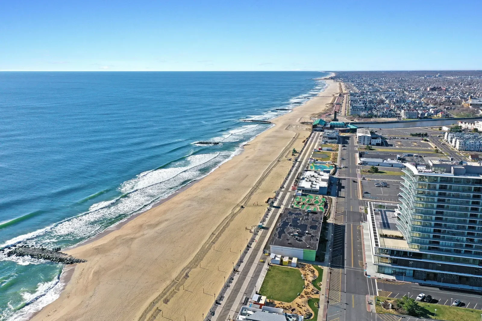 Aerial view of a coastal city showing a sandy beach, ocean waves, and a city skyline with modern buildings and streets.