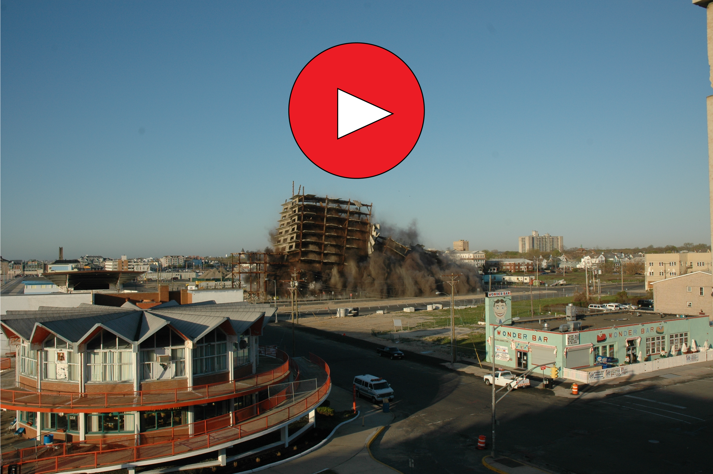 A building collapsing with smoke and debris, on fire, smoke rising into the sky, with commercial buildings and a clear sky in the background.