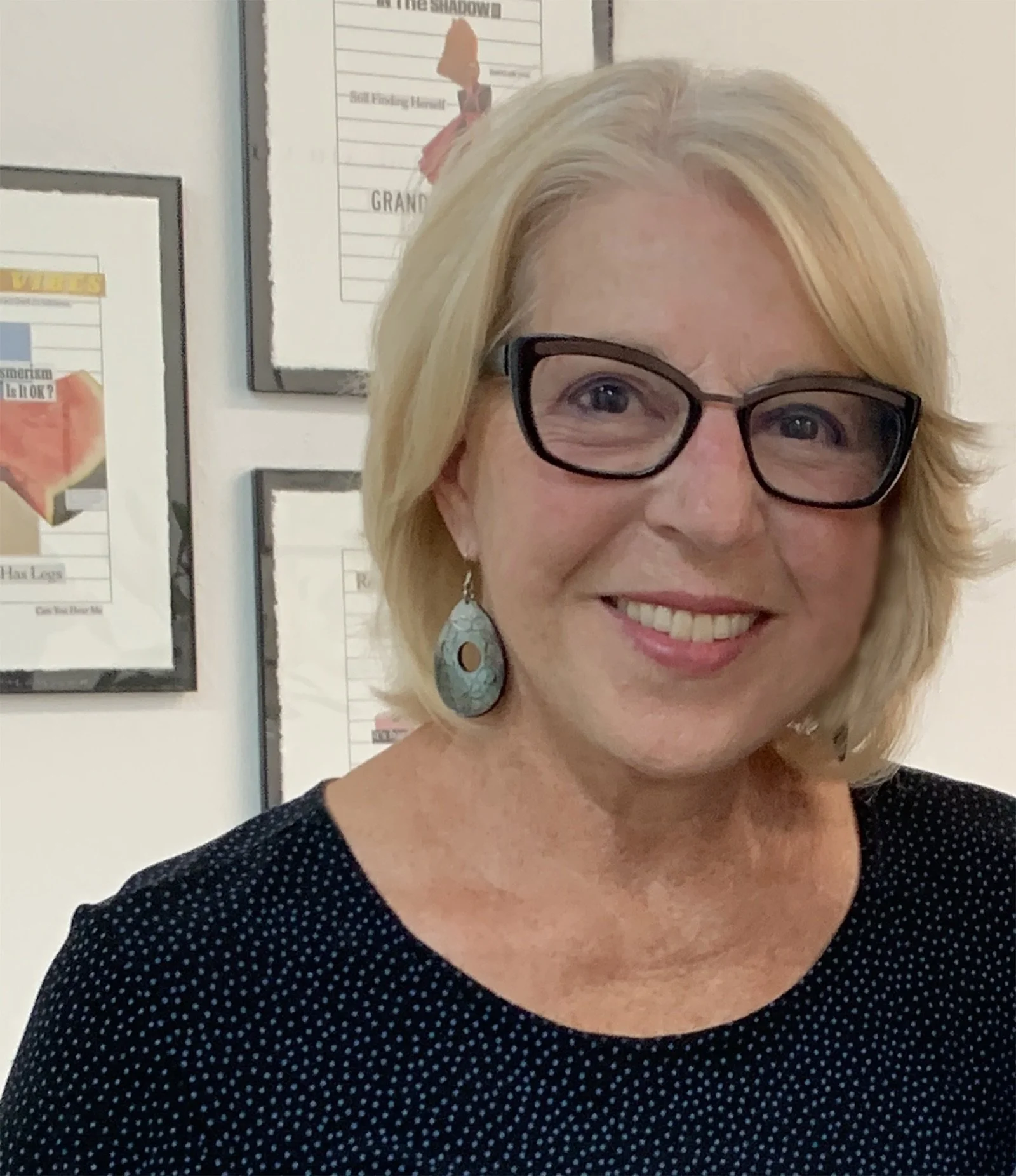 A smiling woman with blonde hair, wearing black glasses and turquoise earrings, standing in front of a wall with framed posters.