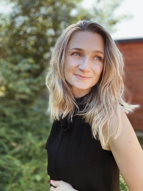 A woman with long blonde hair, wearing a black top, smiling outdoors with trees and a brick wall in the background.