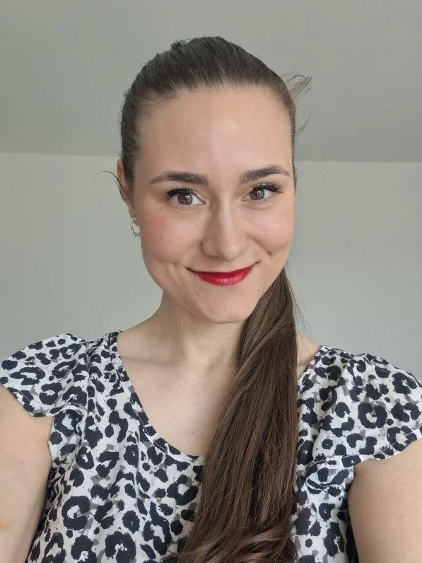 A young woman with fair skin, brown hair tied back, wearing a black and white animal print top, red lipstick, and small earrings, smiling in front of a plain light-colored wall.